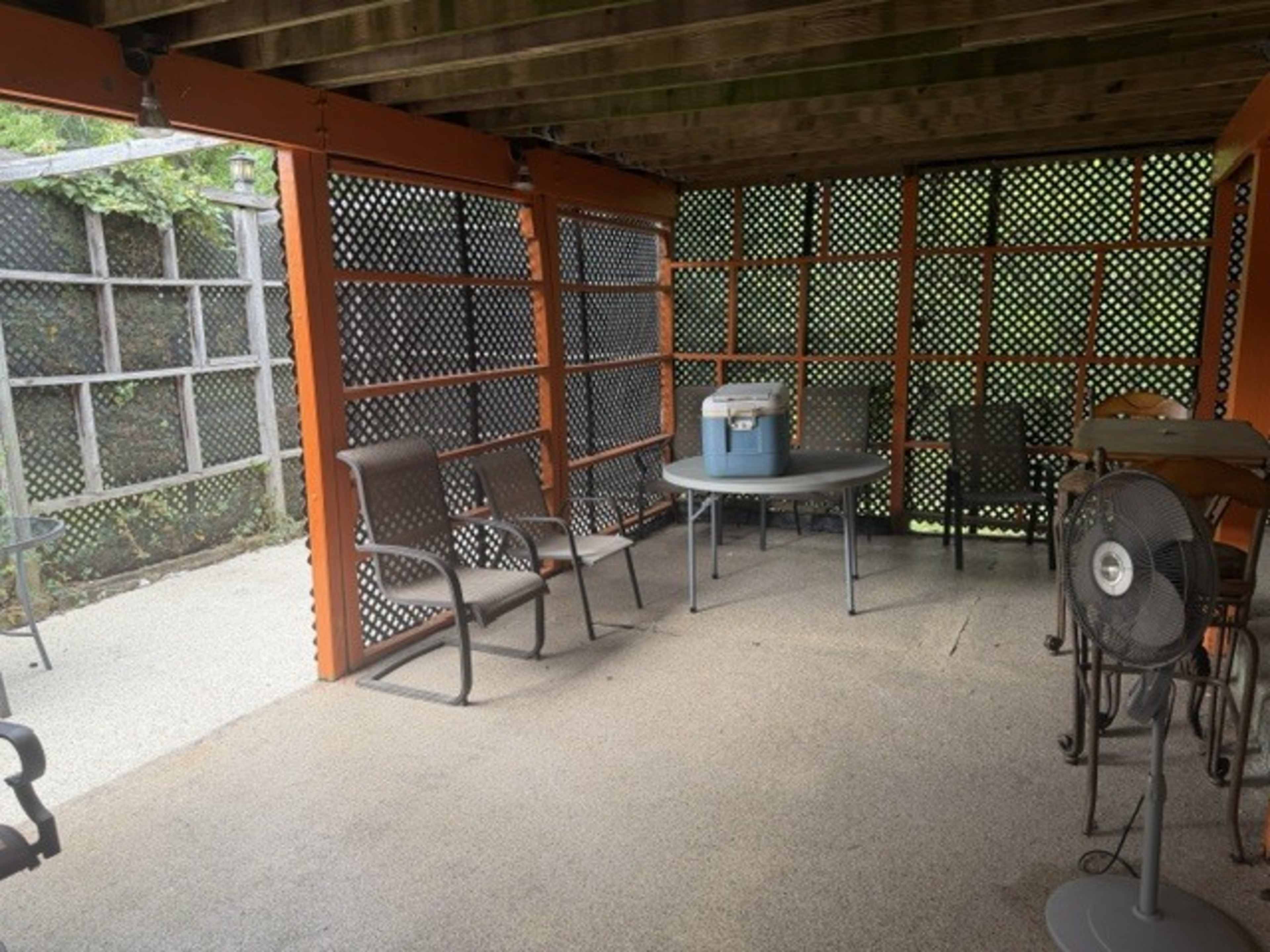 The image shows a covered outdoor patio space with a table, chairs, and a cooler.
