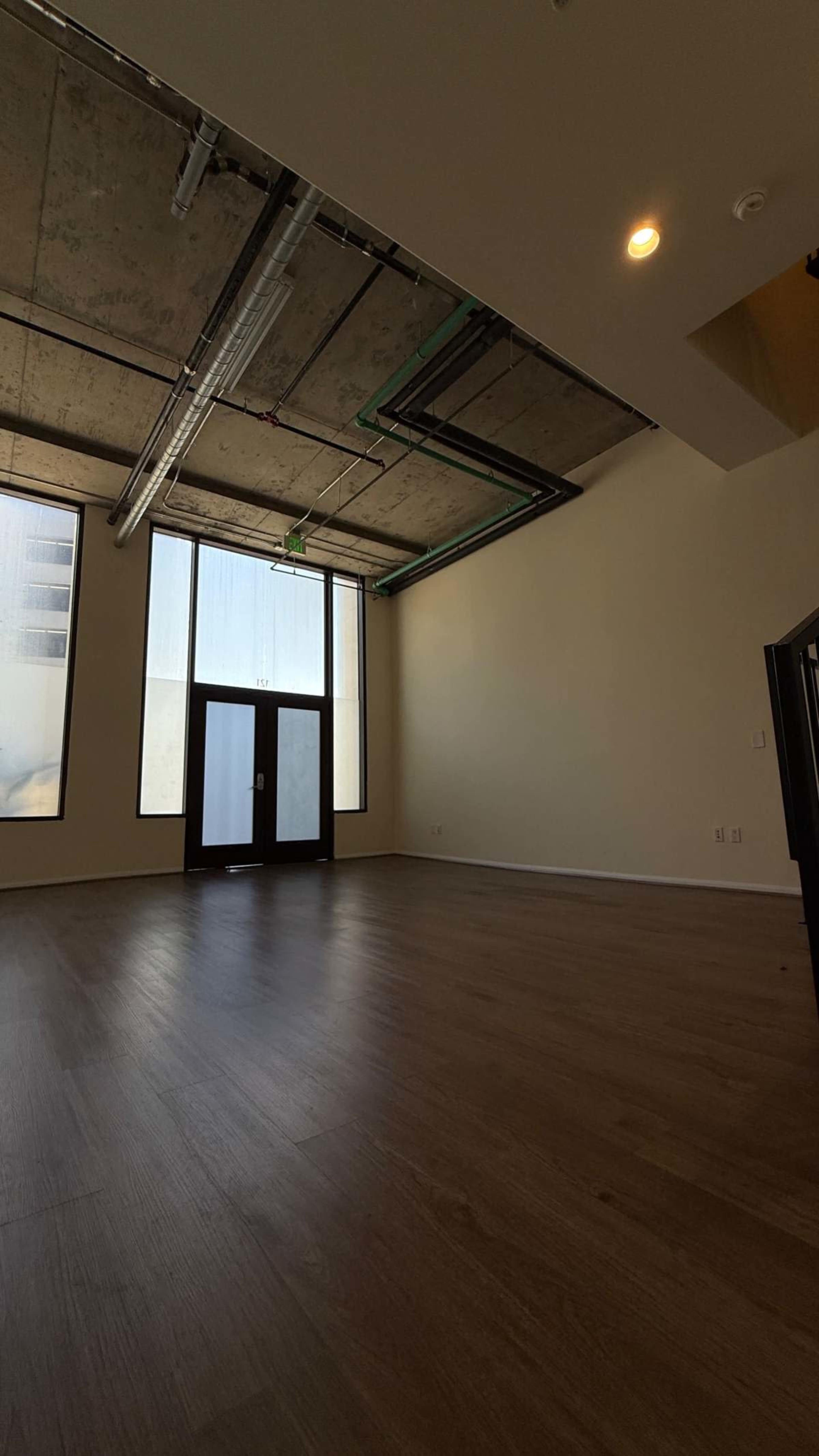 The image shows an empty room with large windows, concrete ceilings, and wooden flooring, featuring a set of double doors at one end.