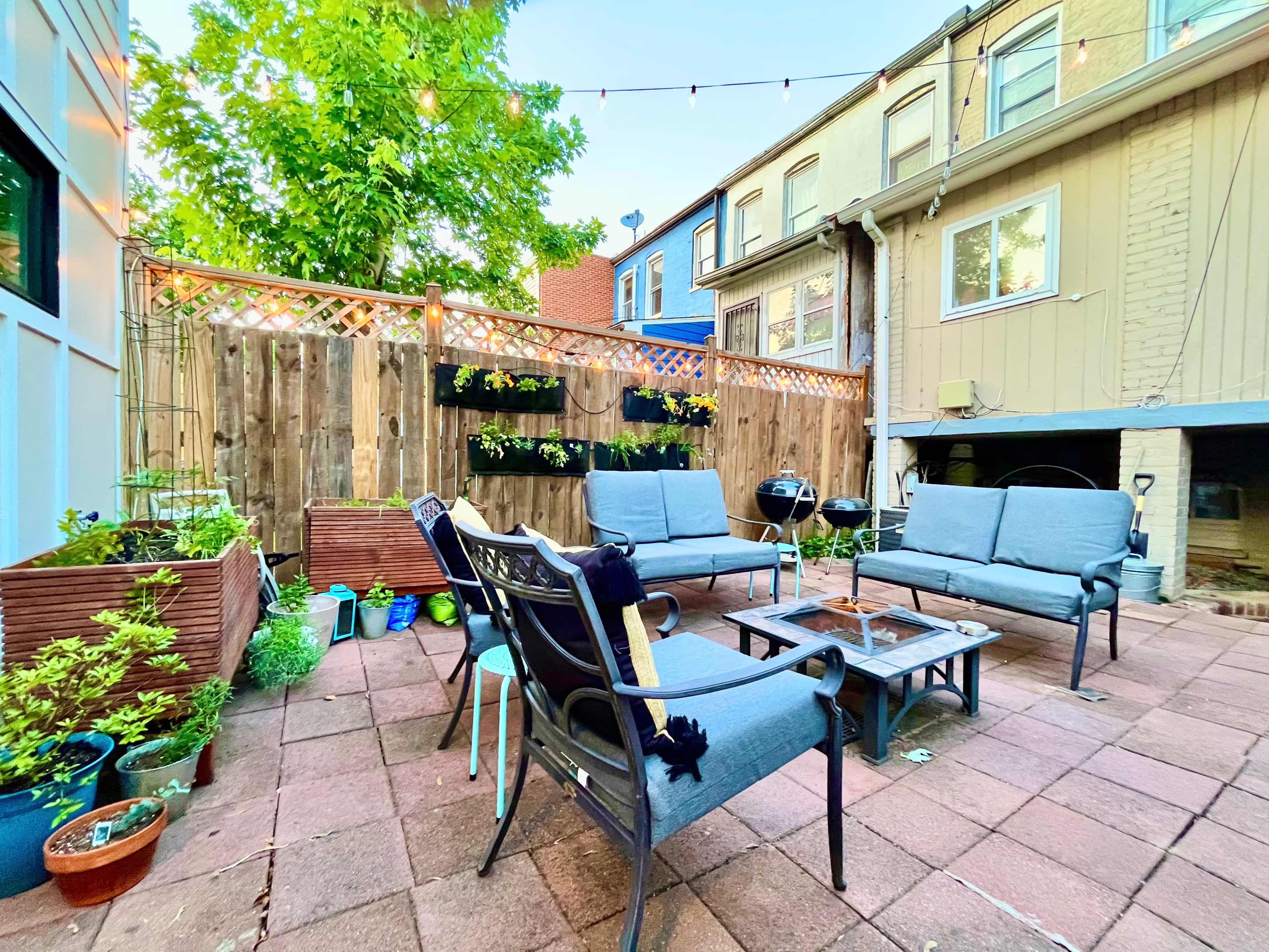 A patio area with gray seating arranged around a glass-top table, surrounded by planters and string lights illuminating a wooden fence.