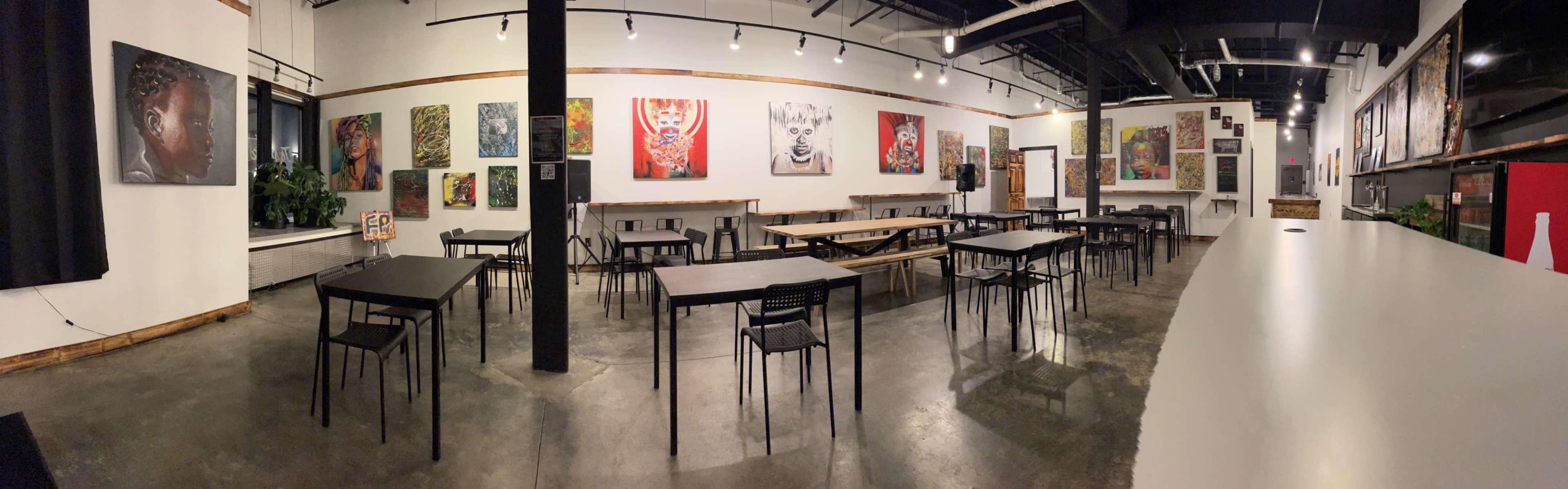 An empty art gallery featuring black tables and chairs arranged around a spacious room adorned with colorful artwork on the walls.