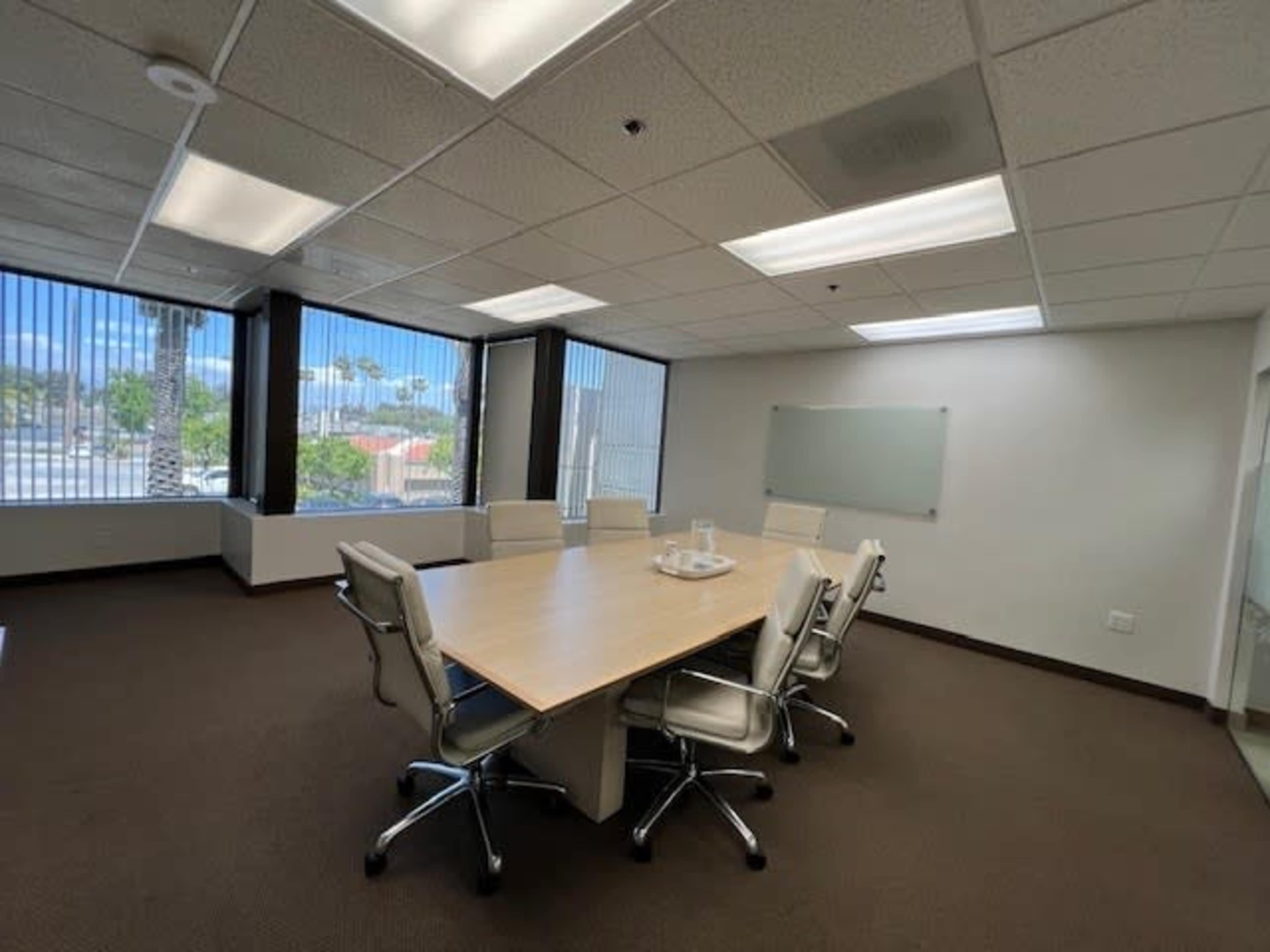 A conference room features a wooden table surrounded by white chairs, with large windows letting in natural light.