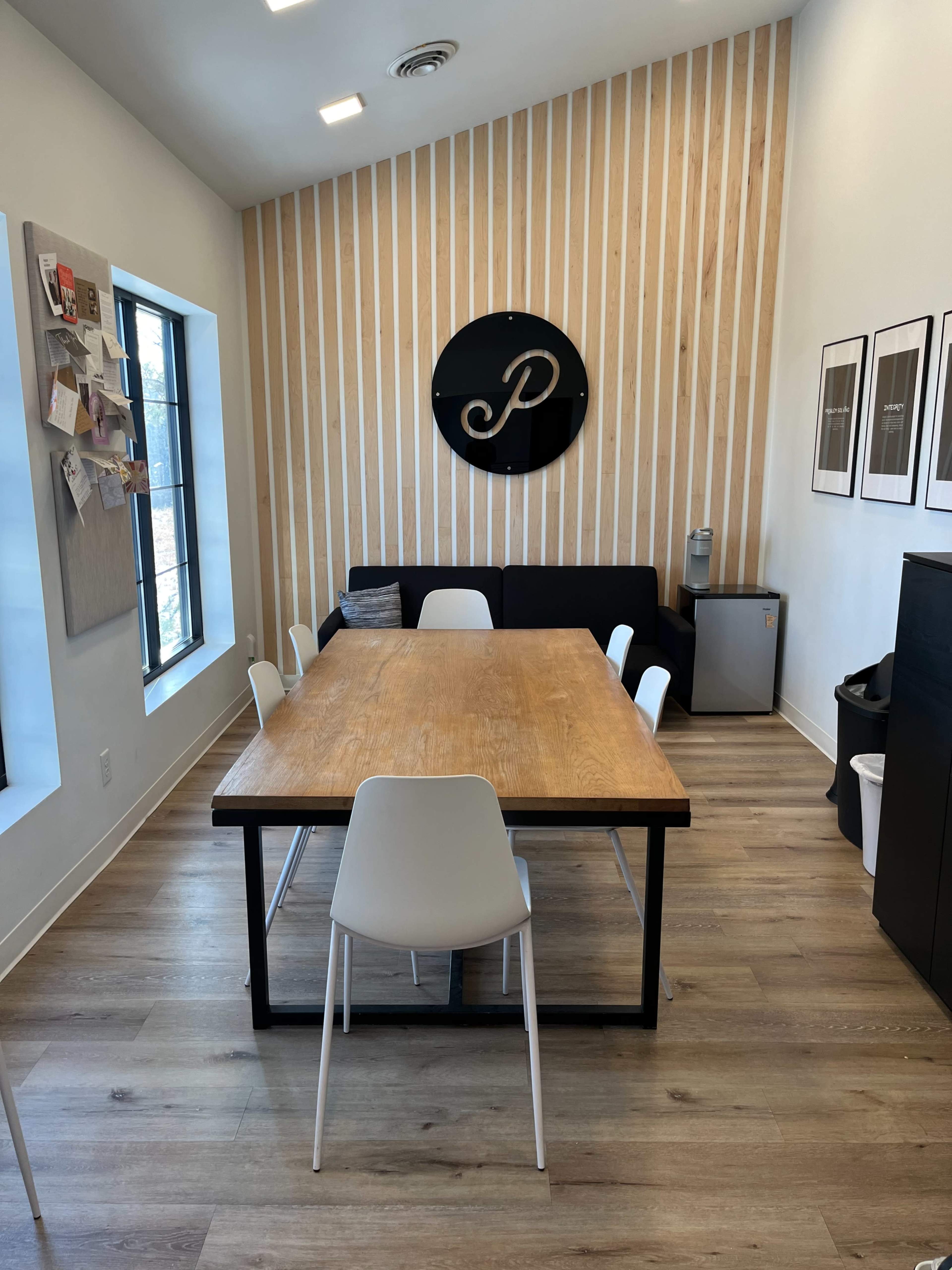 A modern meeting room with a wooden table surrounded by white chairs, a black couch, and a wall featuring wooden paneling and framed pictures.