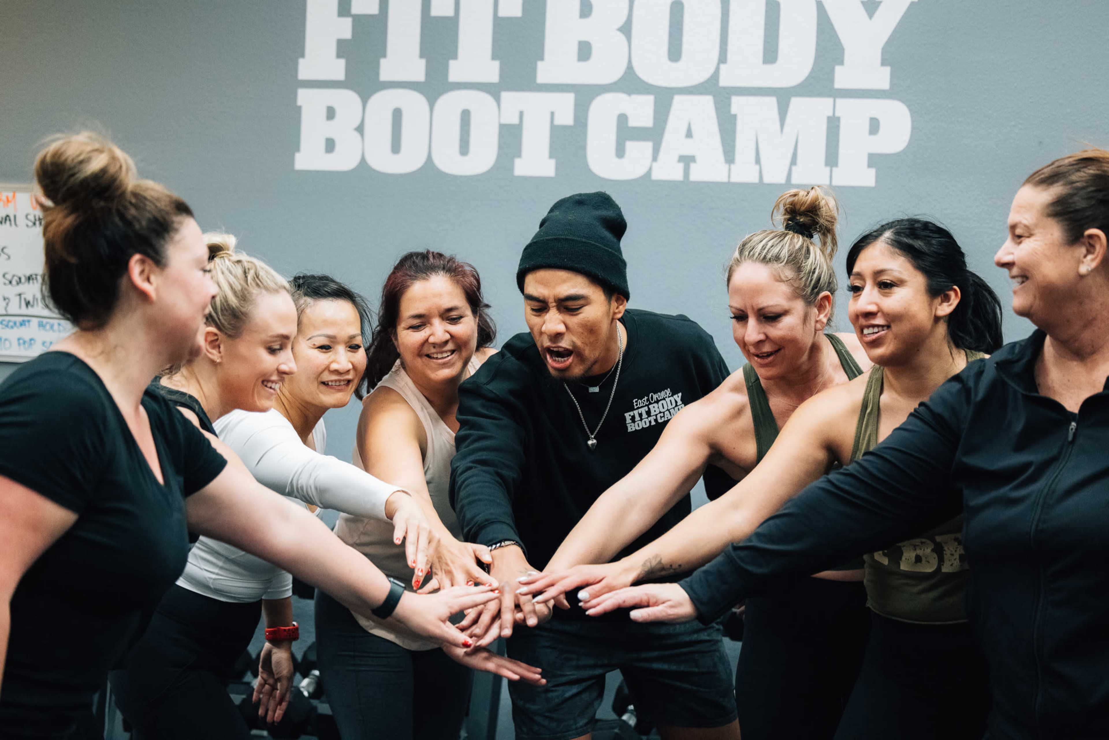 A group of women and a man are standing in a circle with their hands stacked together, showcasing team spirit at a fitness boot camp.