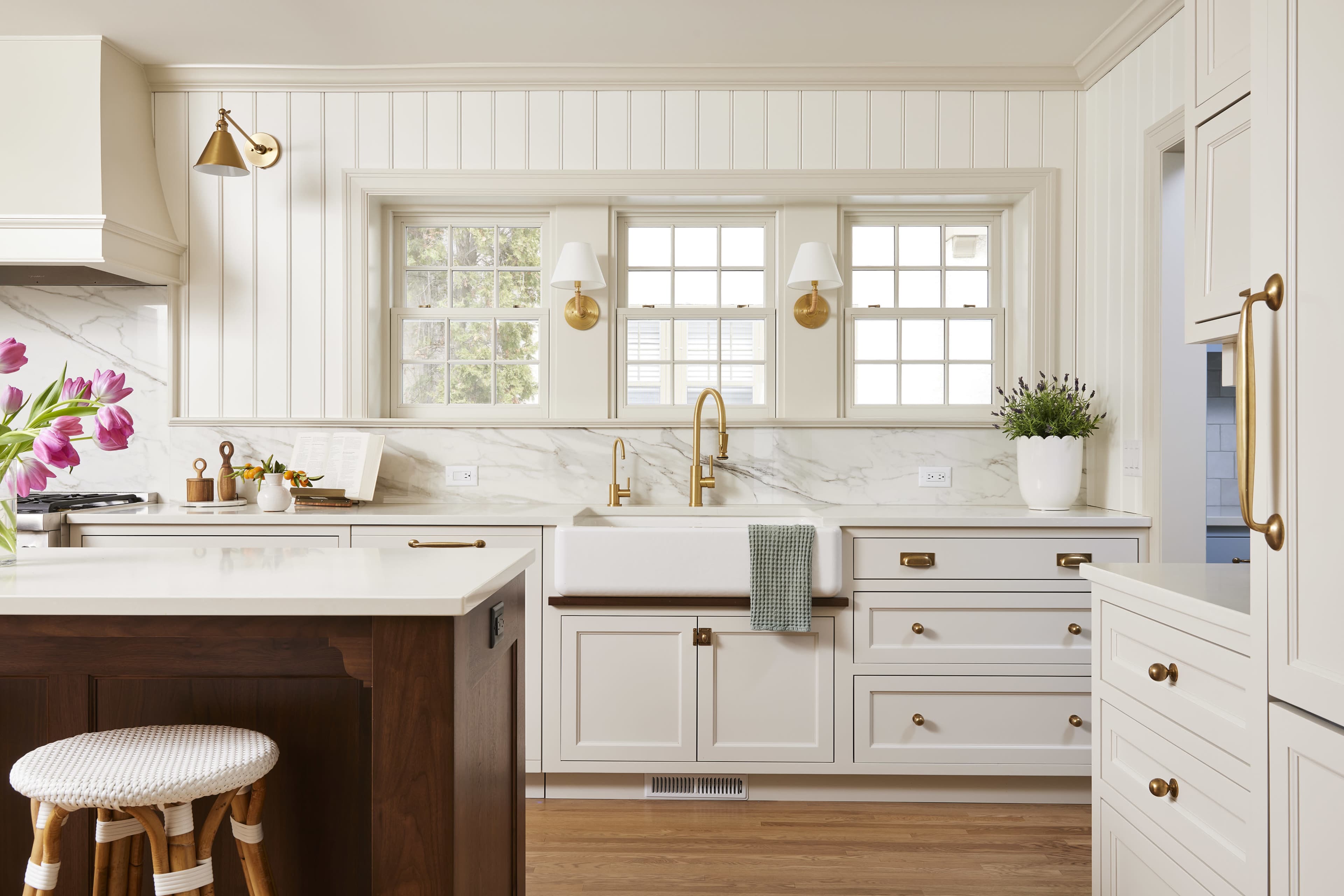 A bright kitchen features white cabinetry, a farmhouse sink, and gold fixtures, with a window view and fresh flowers on the counter.