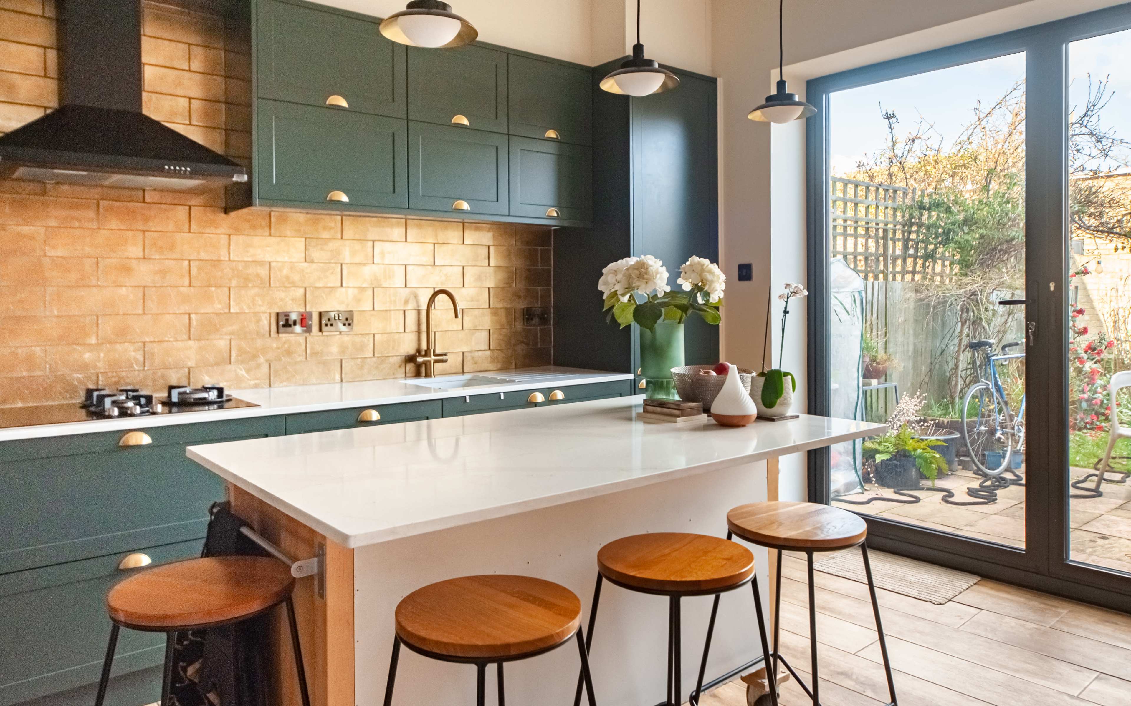 A modern kitchen features a marble island with wooden stools, dark green cabinetry, and a large glass door that leads to an outdoor area.