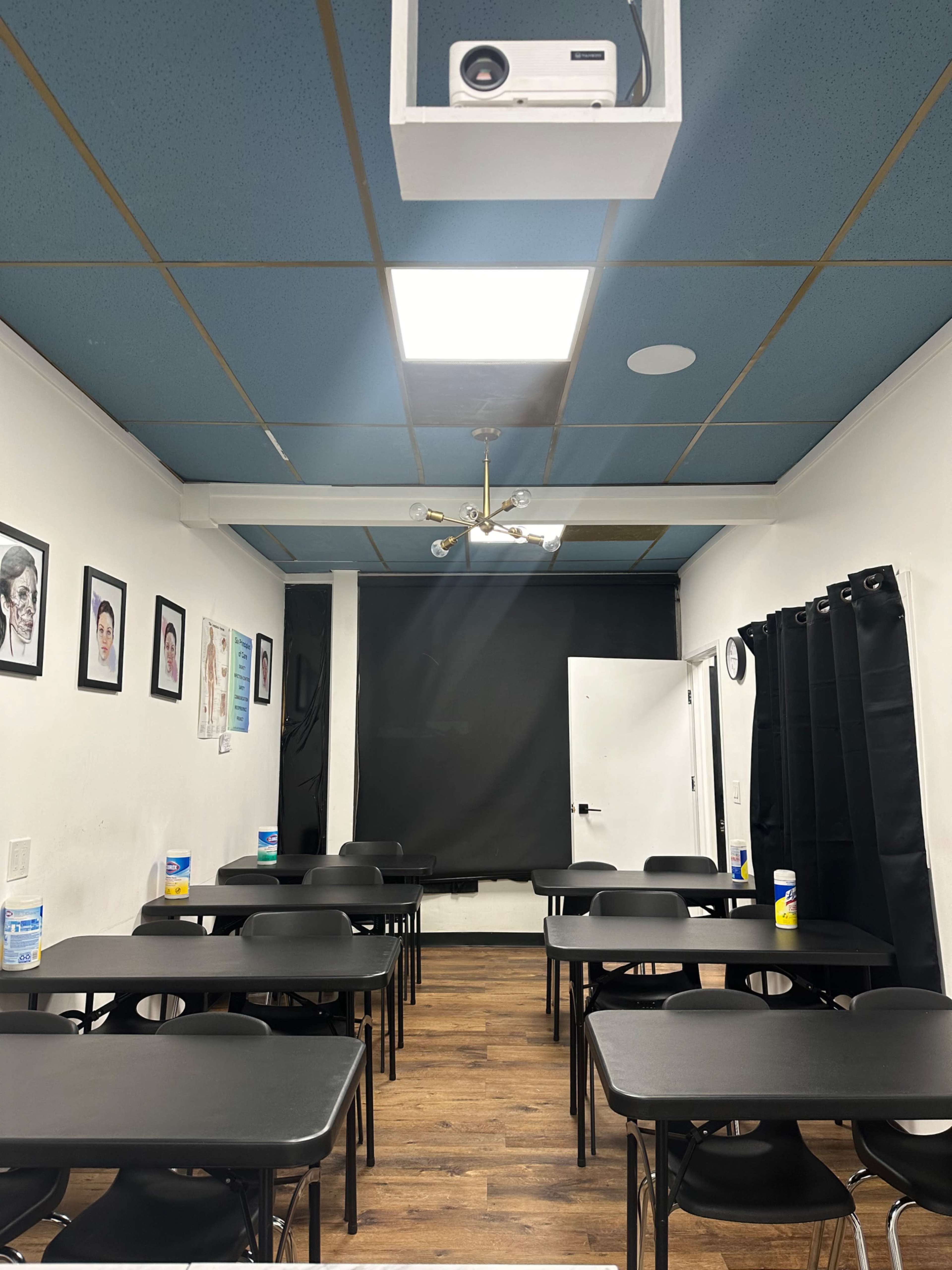 A classroom setup with multiple black tables and chairs, a projector mounted on the ceiling, and black curtains covering a window.