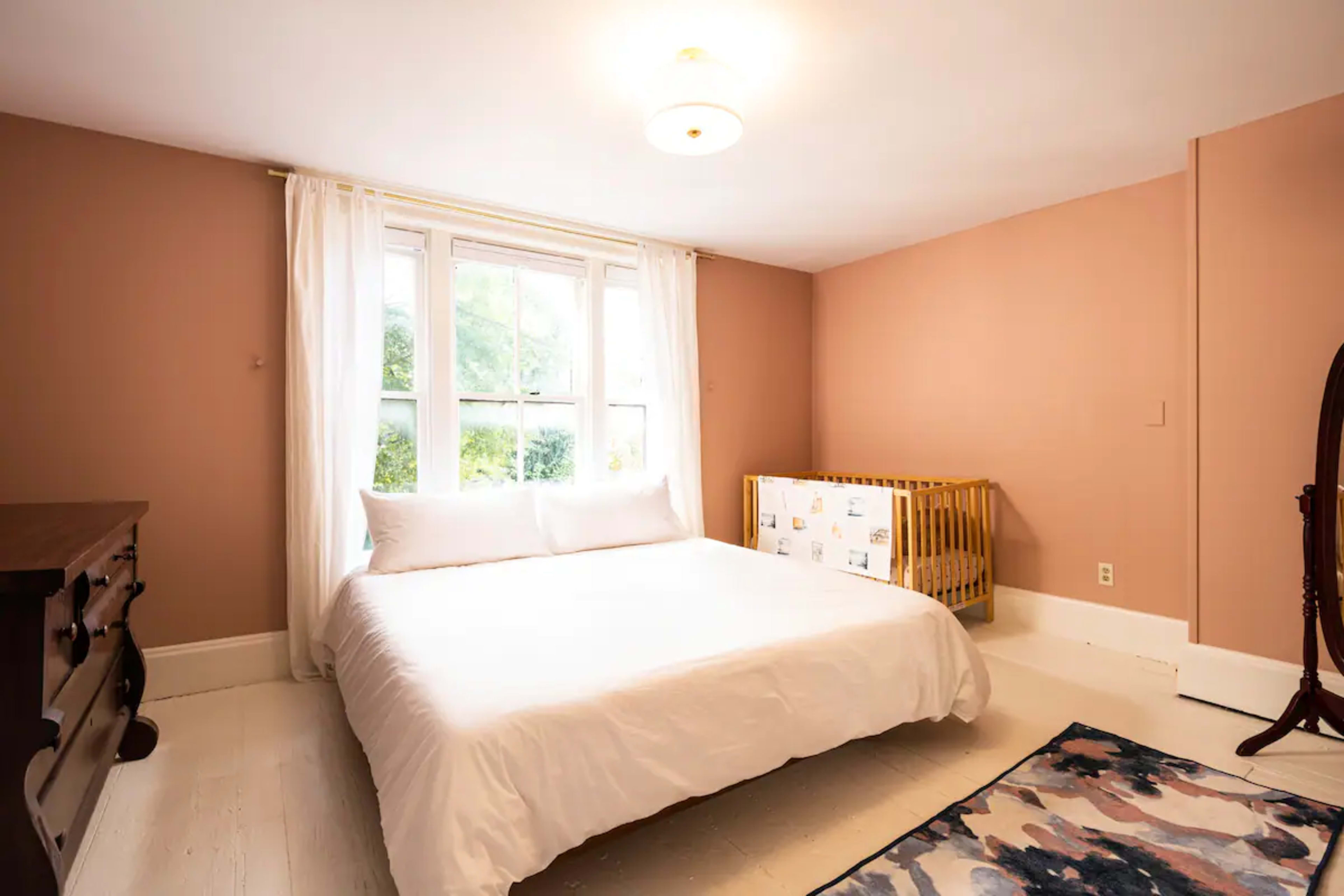 A bedroom with a double bed, a crib, a large window, and a dresser against the wall.