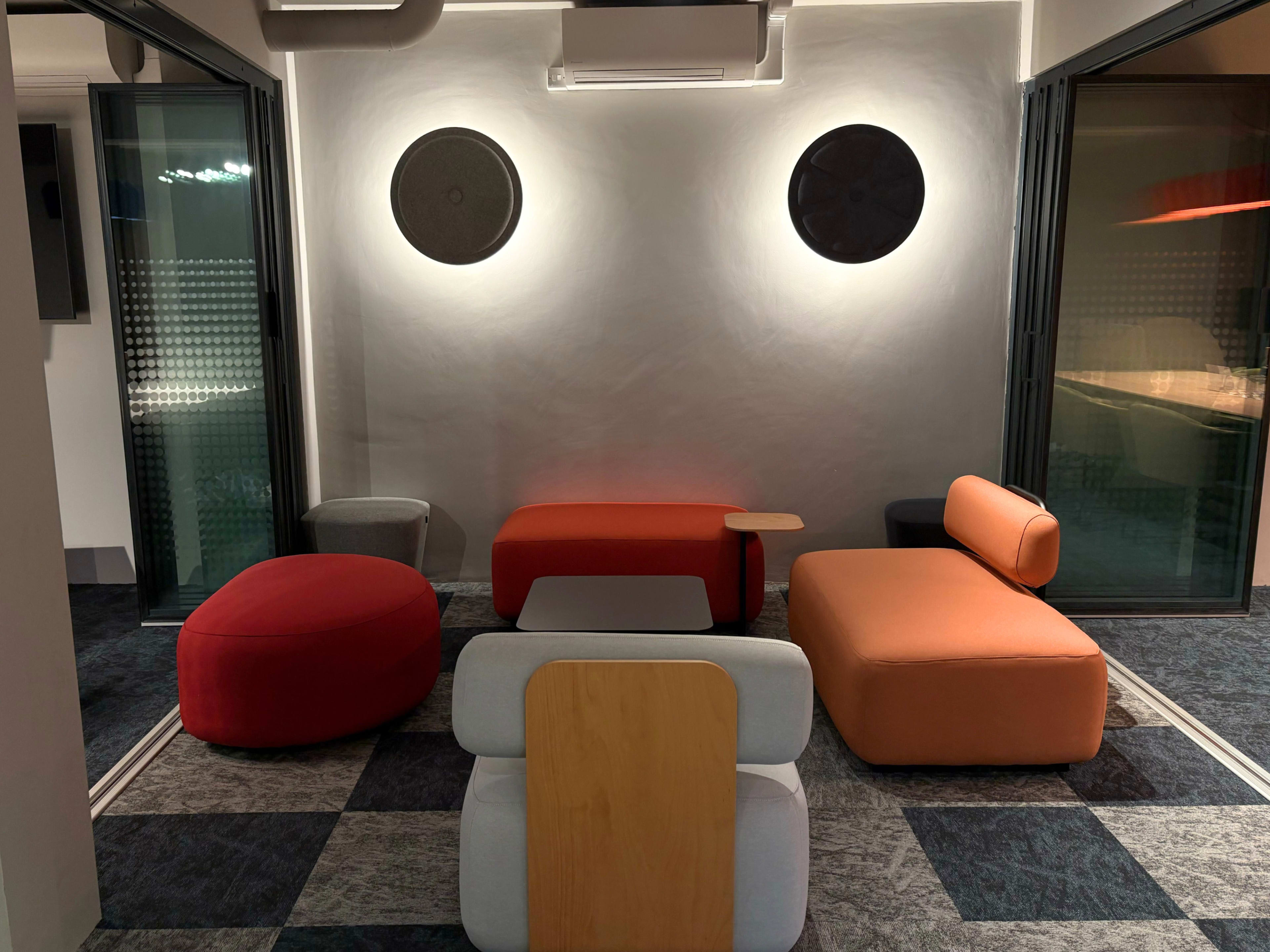 The image shows a modern lounge area with various upholstered seating options in red, gray, and orange, arranged around a light-colored side table.