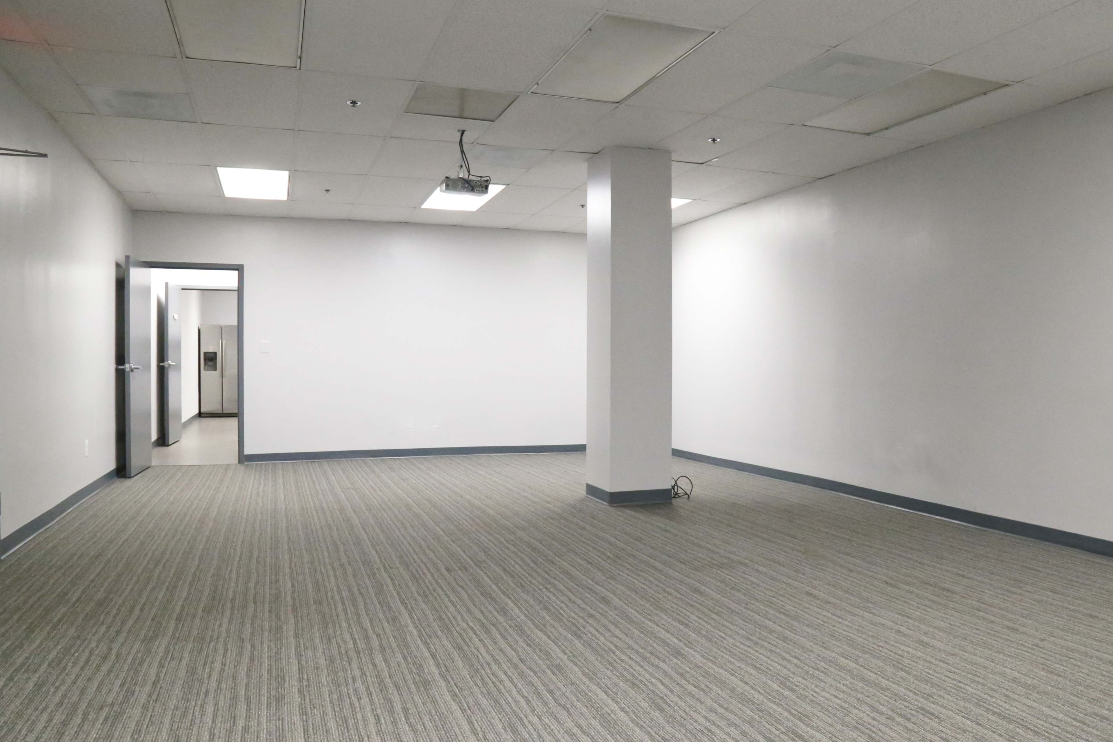 A vacant office space with gray carpet, white walls, and a single central column.