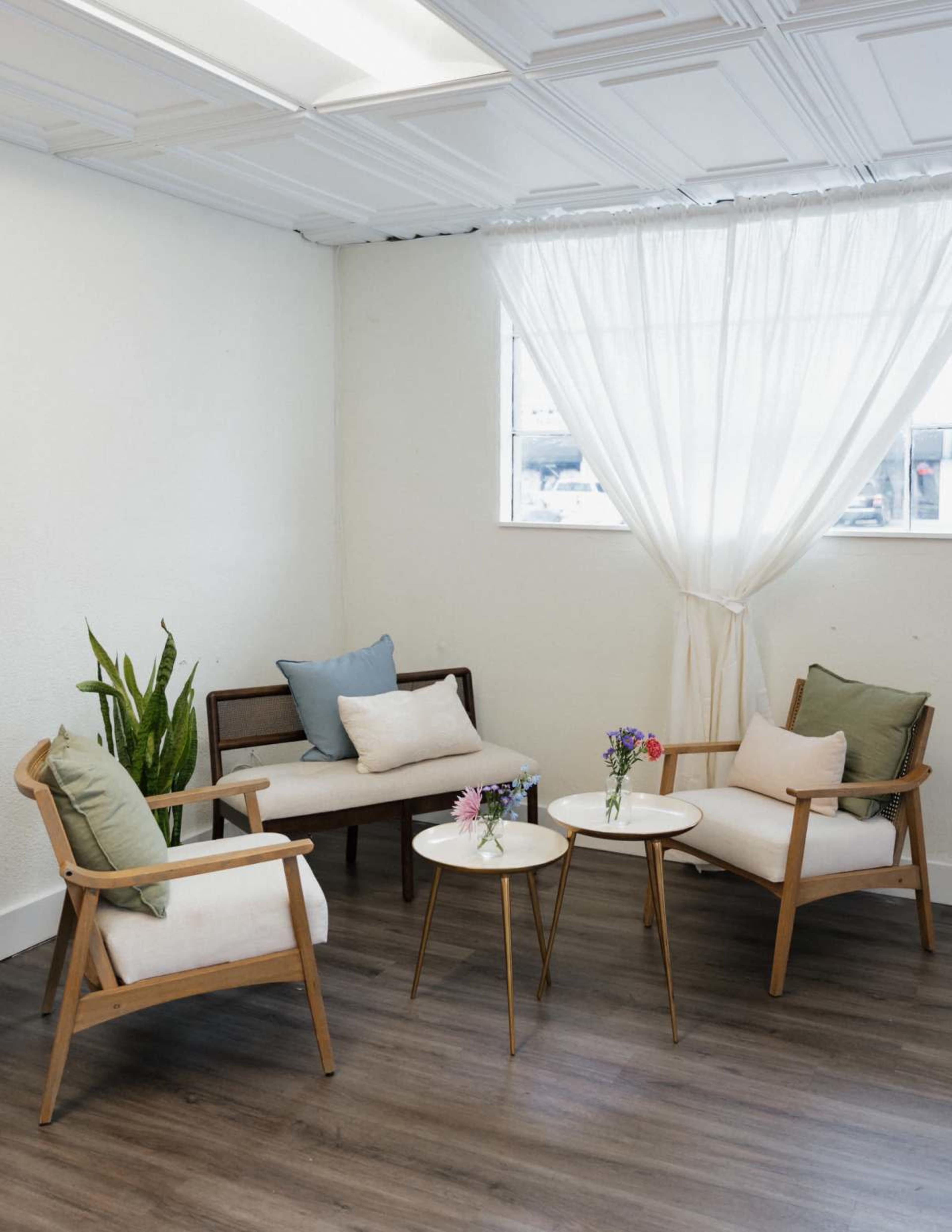The image shows a minimalist seating area with two wooden chairs and a sofa, accompanied by small round tables and a large window covered by sheer curtains.