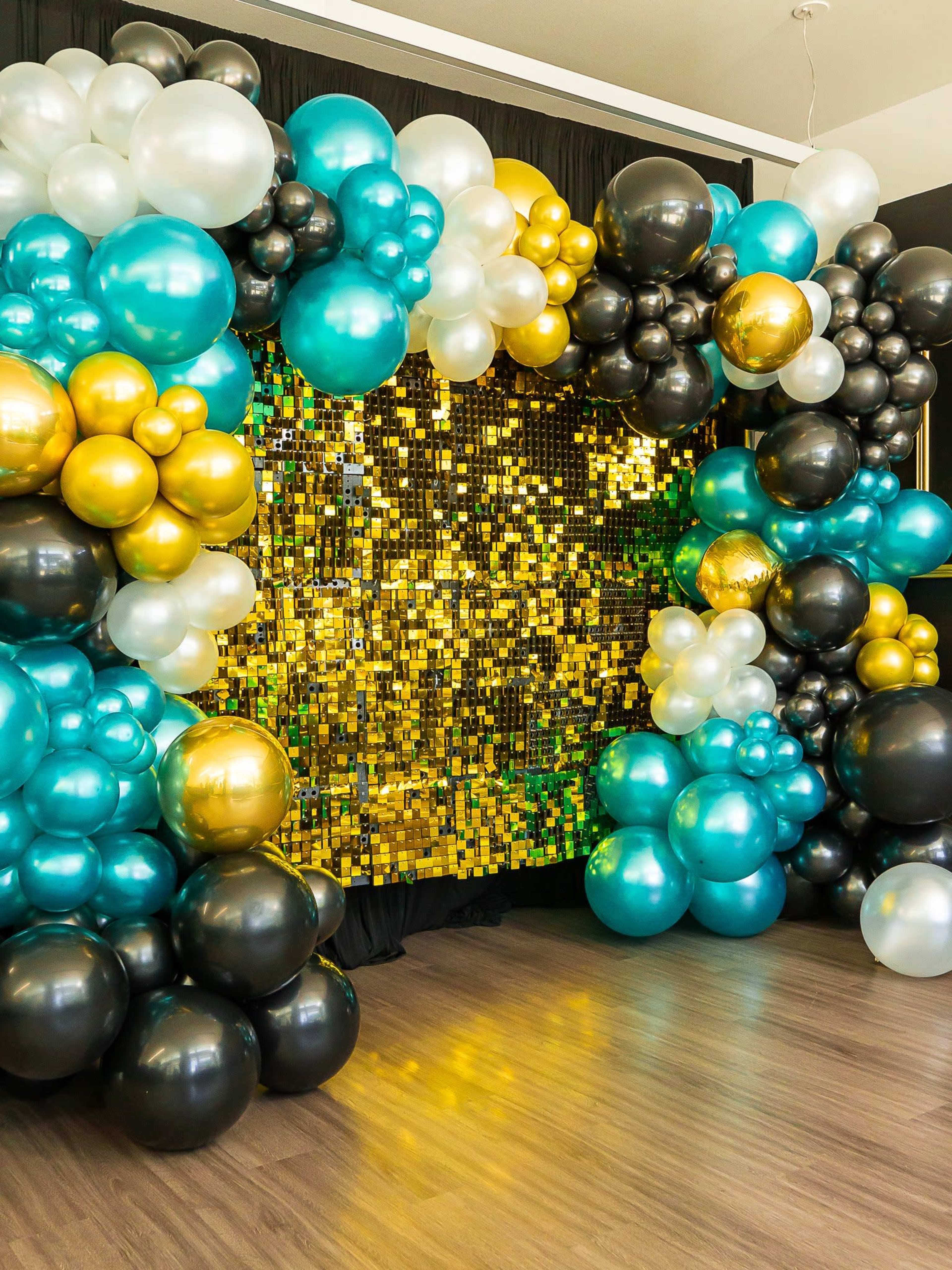 The image features a decorative backdrop adorned with an array of balloons in black, gold, and teal, surrounding a shimmering gold sequin wall.
