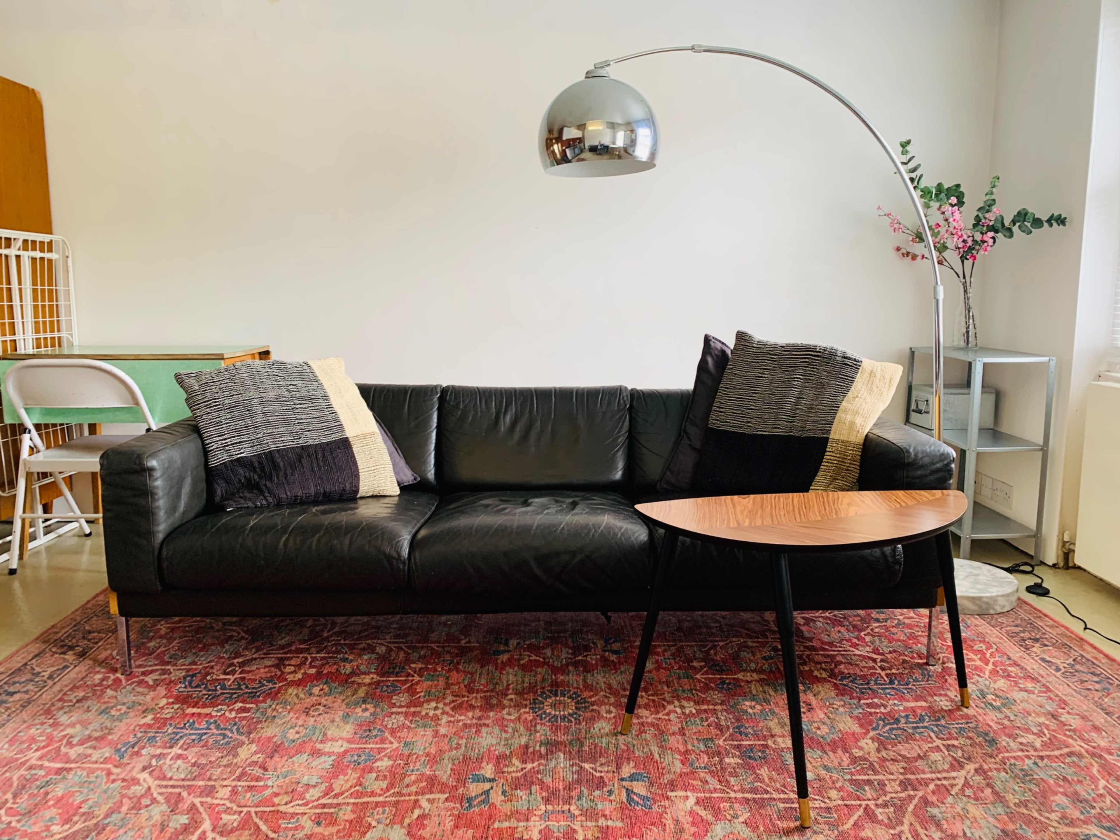 A black leather sofa with two textured cushions is positioned next to a curved metallic floor lamp and a small wooden coffee table on a colorful rug.