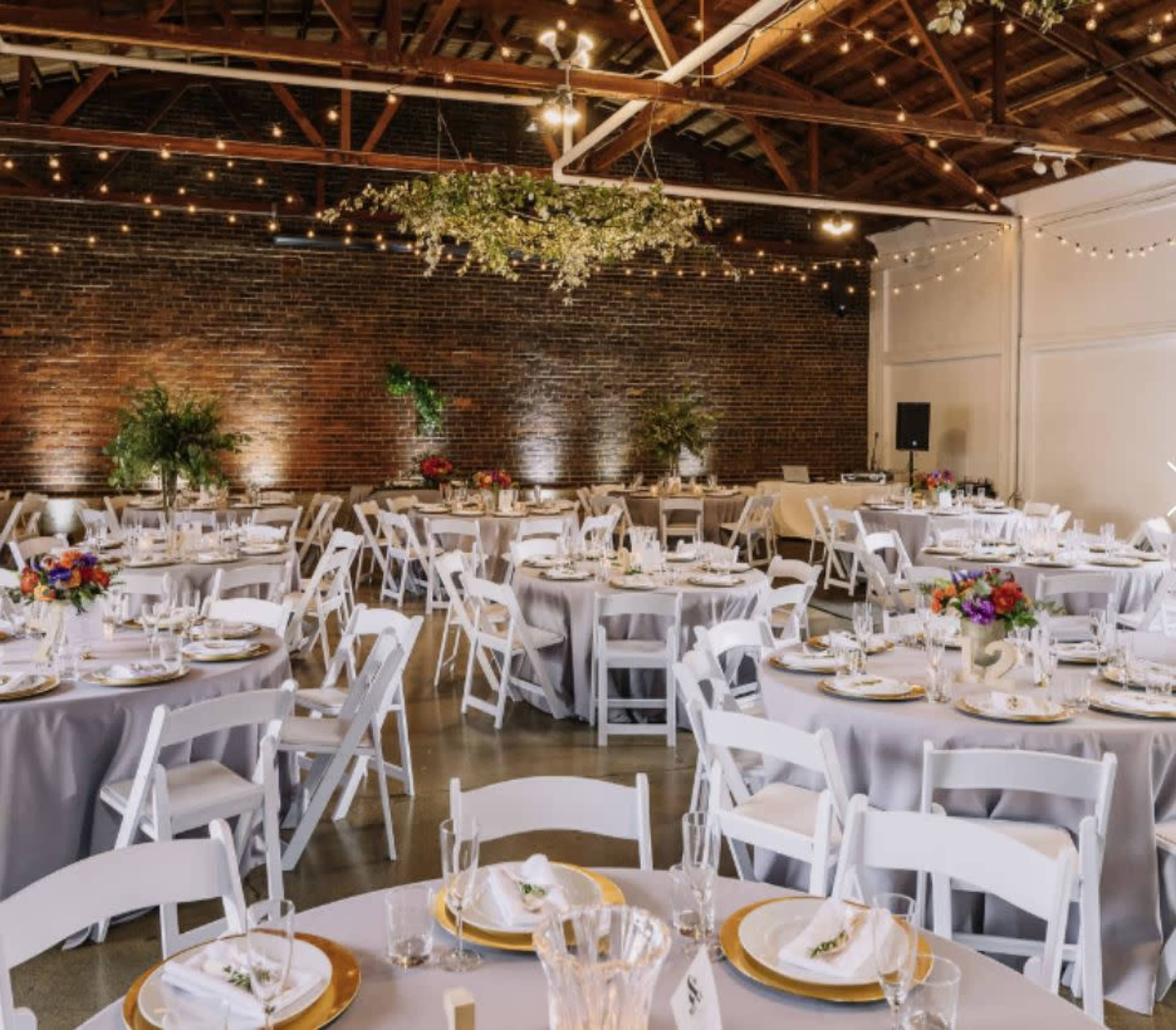 A spacious event venue features round tables set with white chairs and gray table linens, adorned with floral centerpieces and overhead string lights.