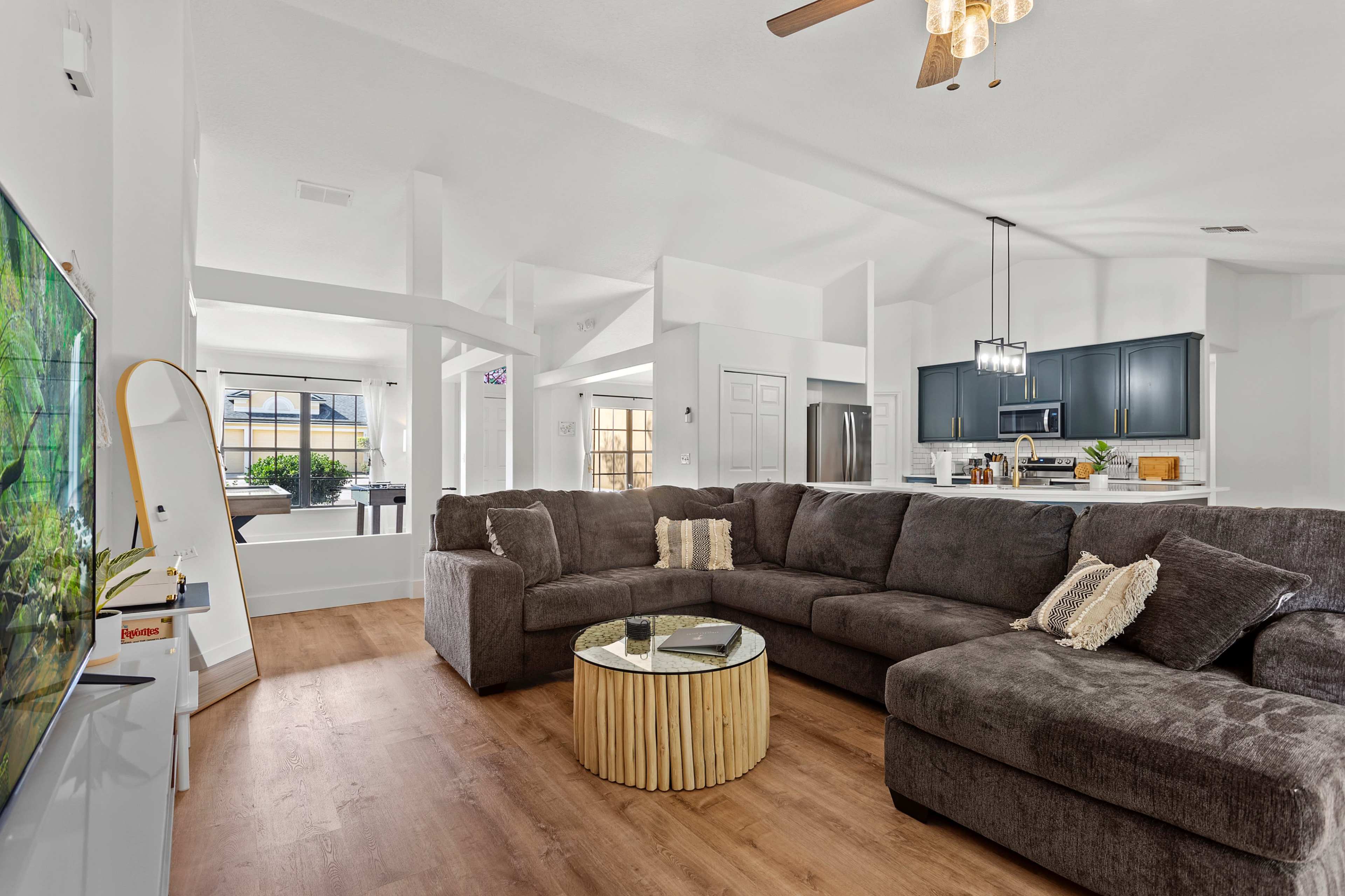 A spacious living room features a large sectional sofa, a round wooden coffee table, and a view of an open kitchen with dark cabinetry.