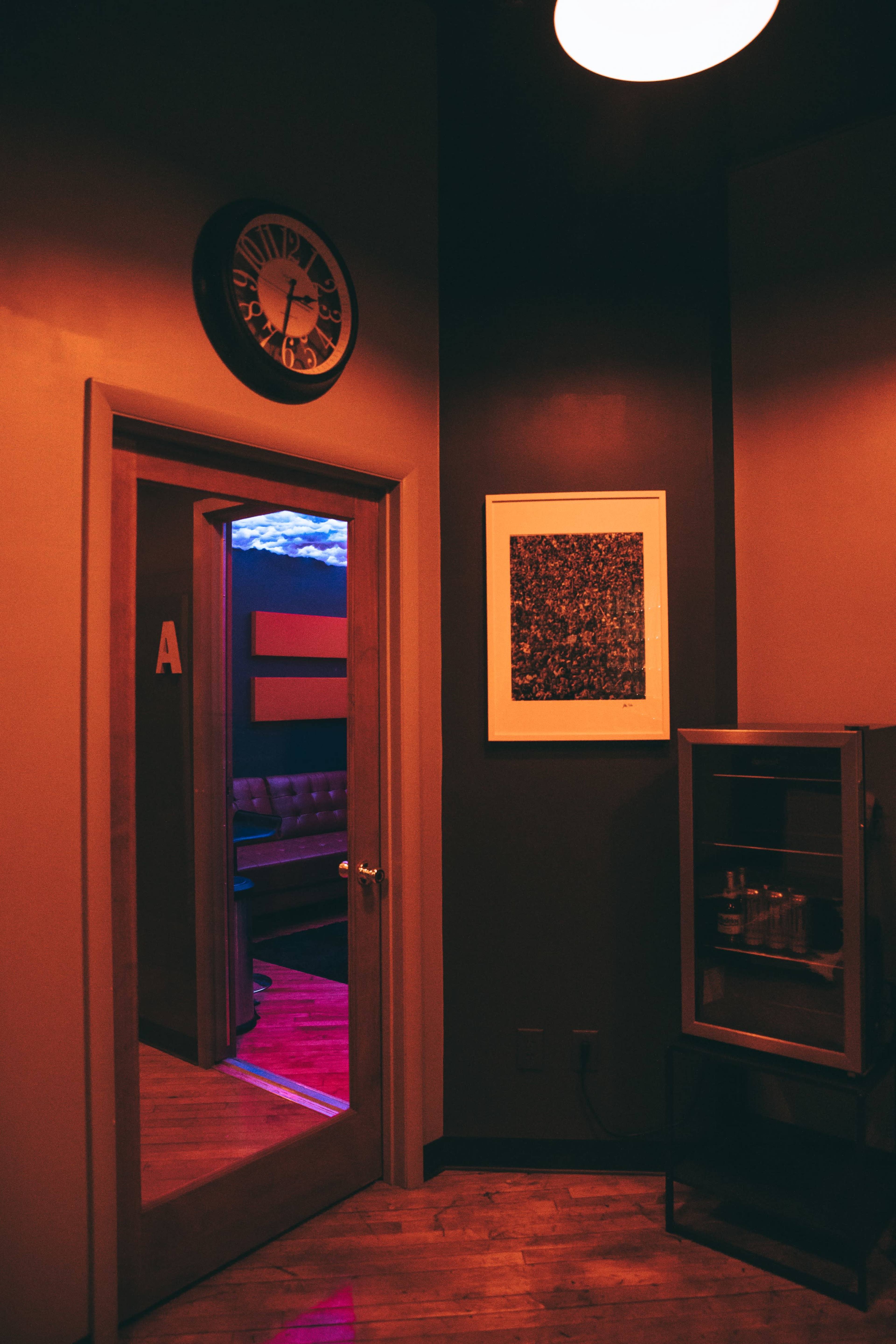 A door with a clock above it leads to a room illuminated by purple light, featuring a patterned artwork on the wall and a wooden cabinet in the corner.