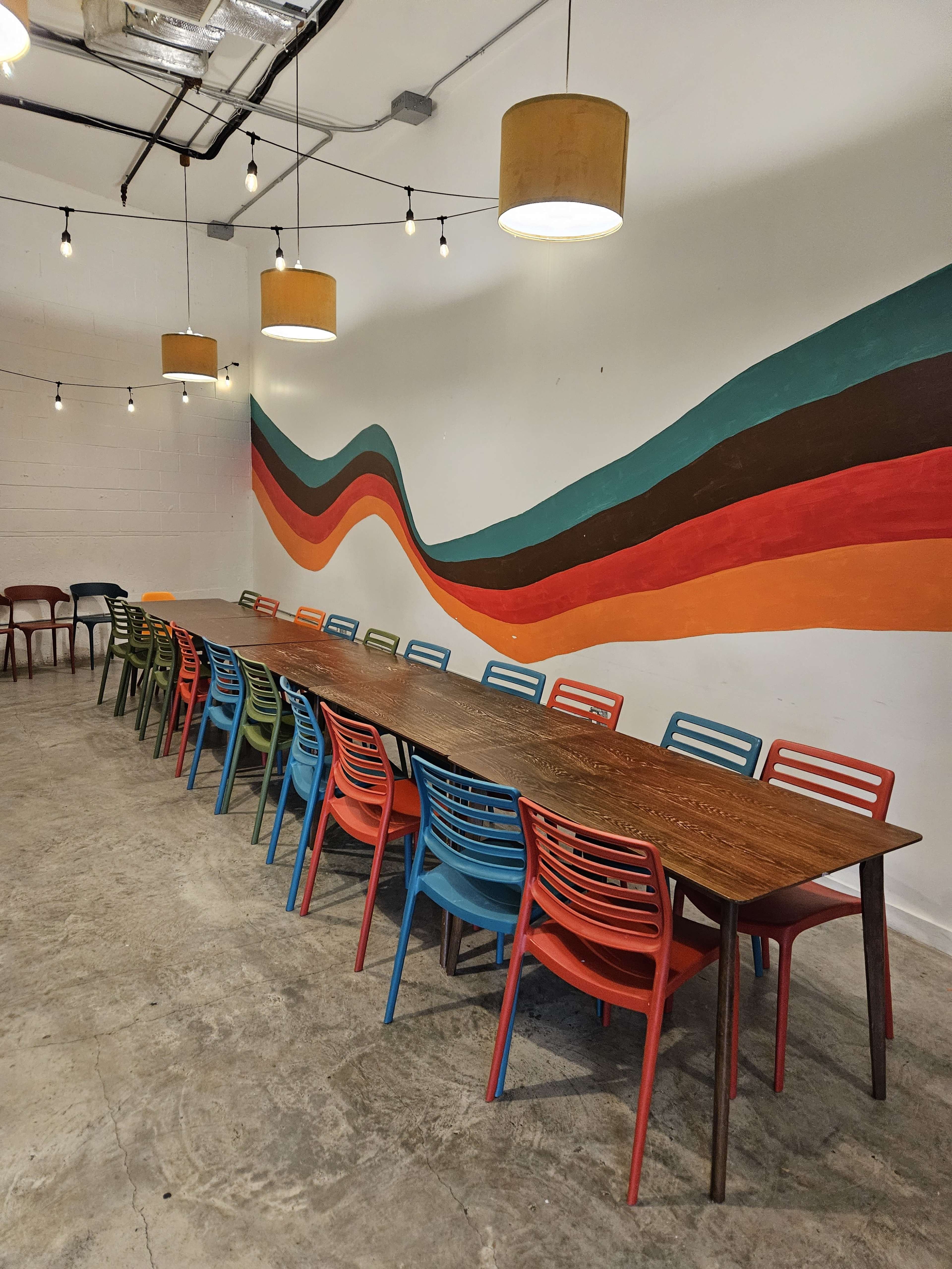 A long wooden table is surrounded by colorful plastic chairs in a room with a striped mural on the wall and pendant lights hanging from the ceiling.
