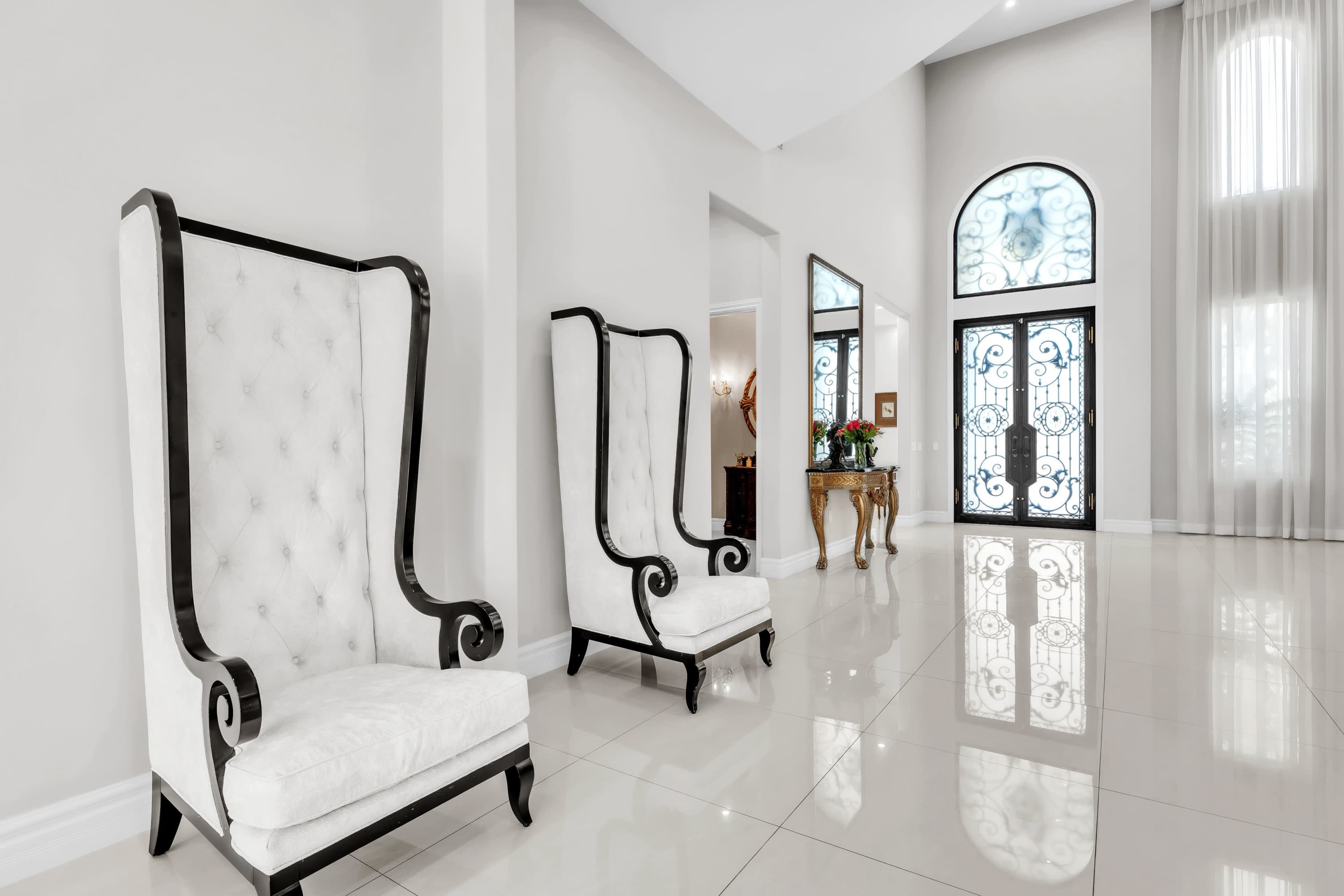 The scene features two white tufted high-back chairs positioned against a light-colored wall, with a large decorative mirror and an arched glass door in the background.
