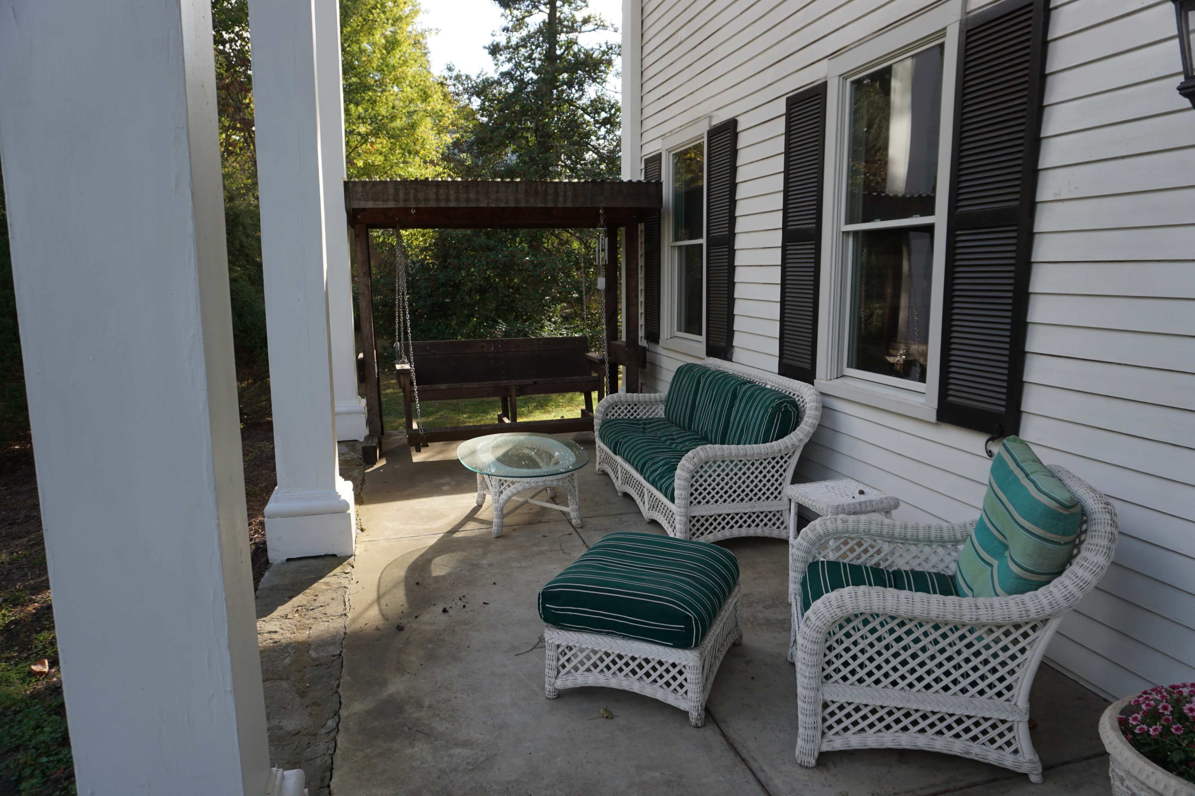 A porch with white wicker furniture, including a sofa, chairs, and a coffee table, alongside a wooden swing and surrounded by greenery.