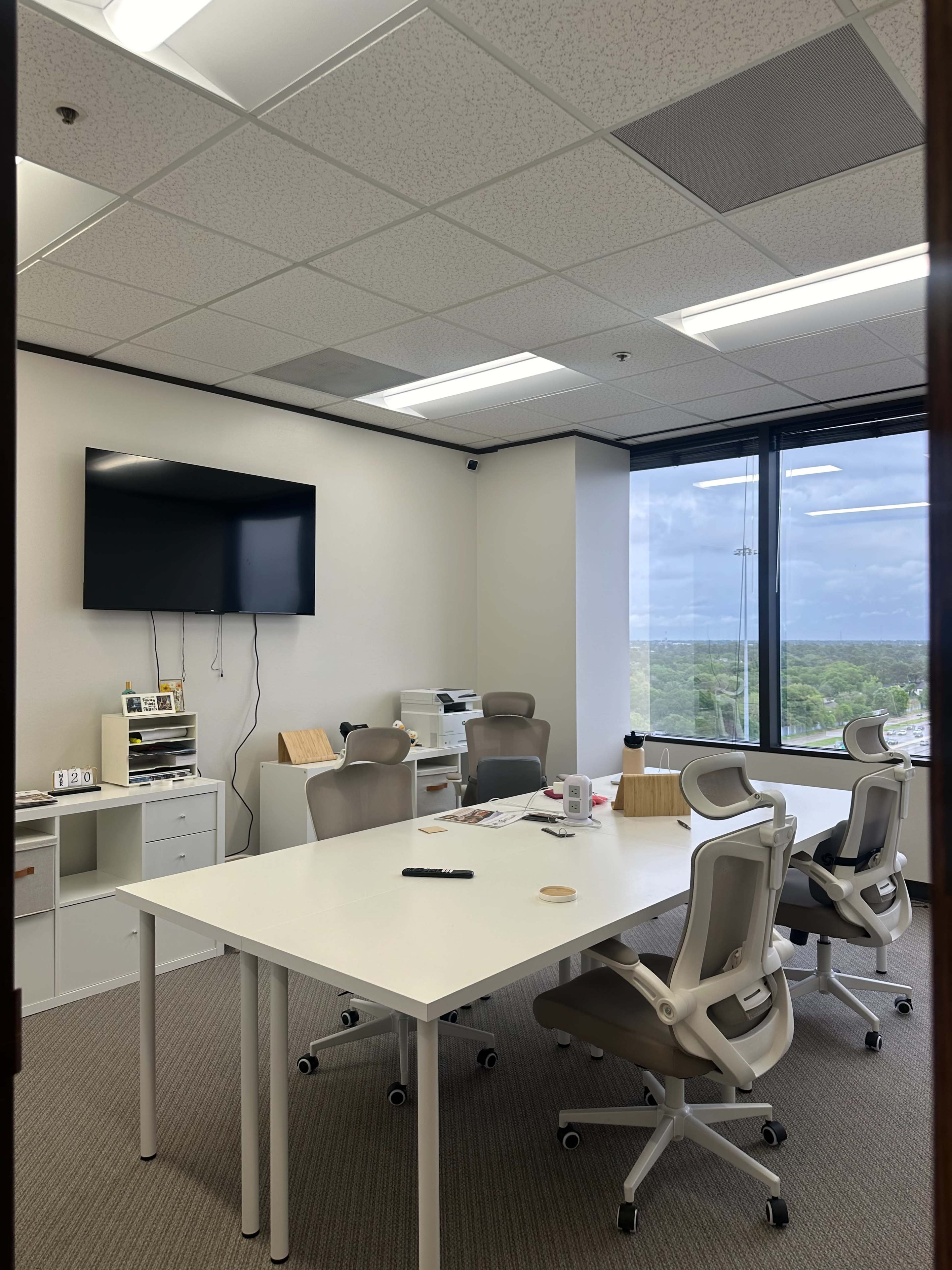 A modern office conference room features a large table surrounded by ergonomic chairs, a wall-mounted TV, and a view of the outdoors through a large window.