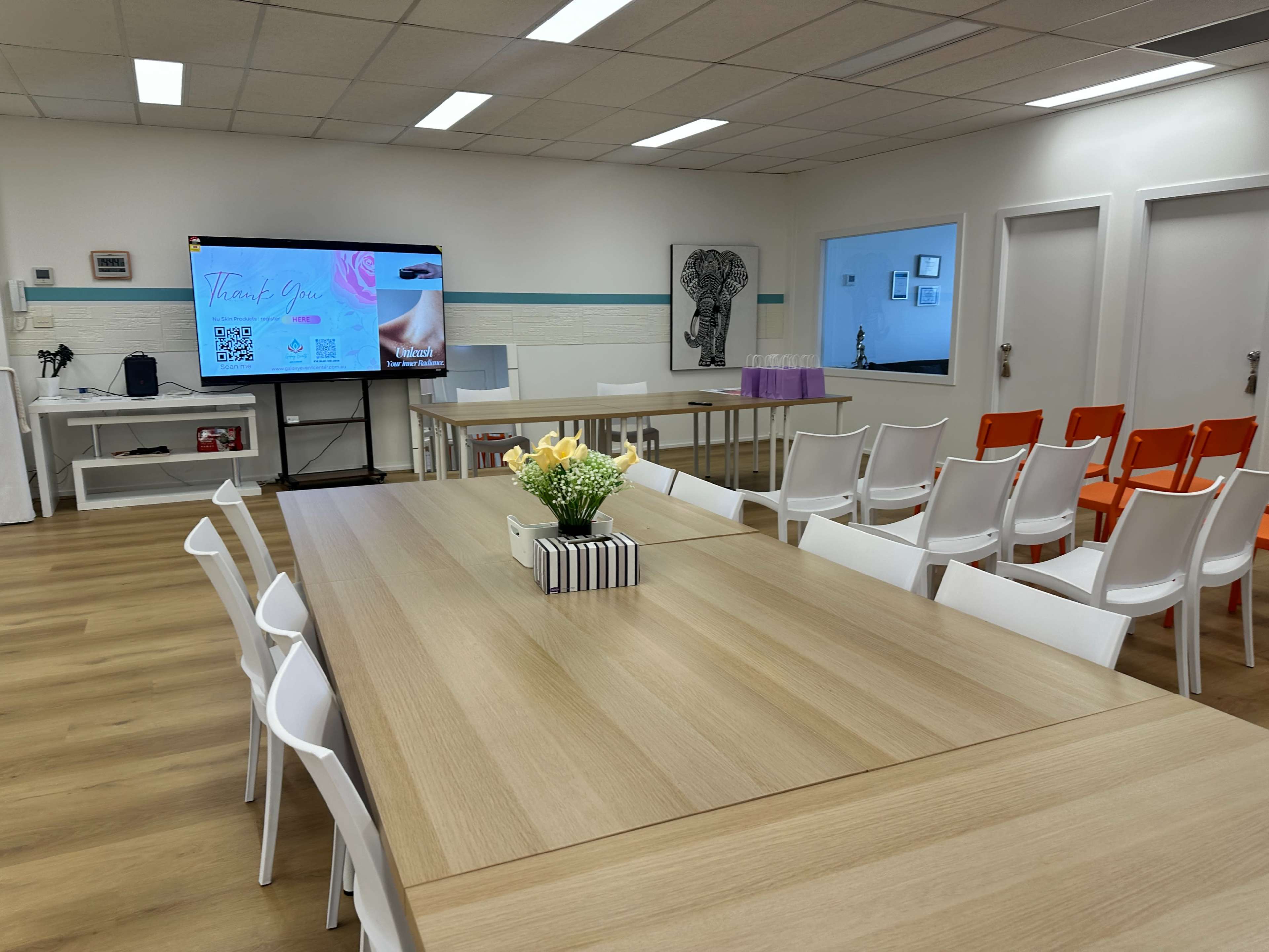 A spacious meeting room features a large table, white chairs, a presentation screen displaying graphics, and a vase of flowers at the center.