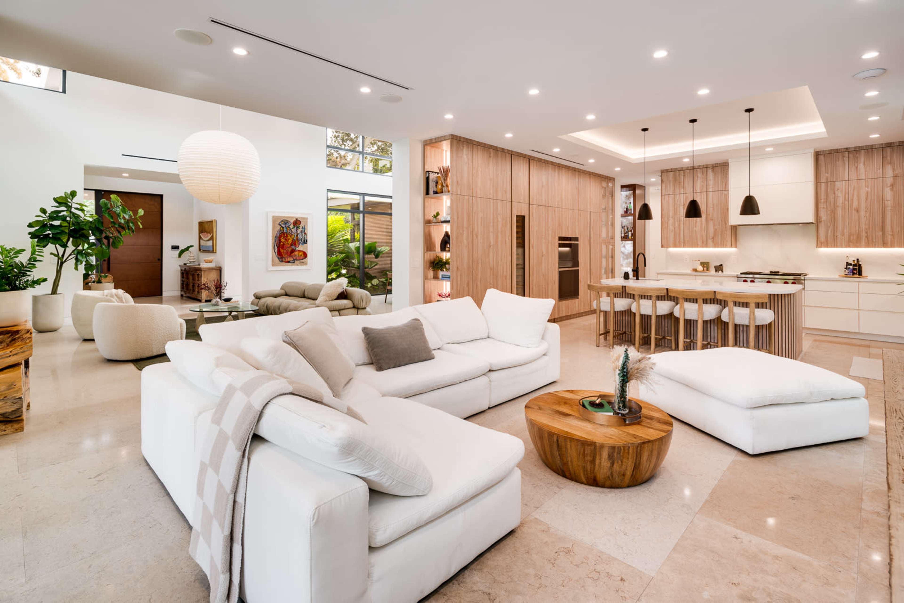 The image shows a modern living room that features a spacious layout with a white sectional sofa, natural wood accents, and a kitchen area in the background.