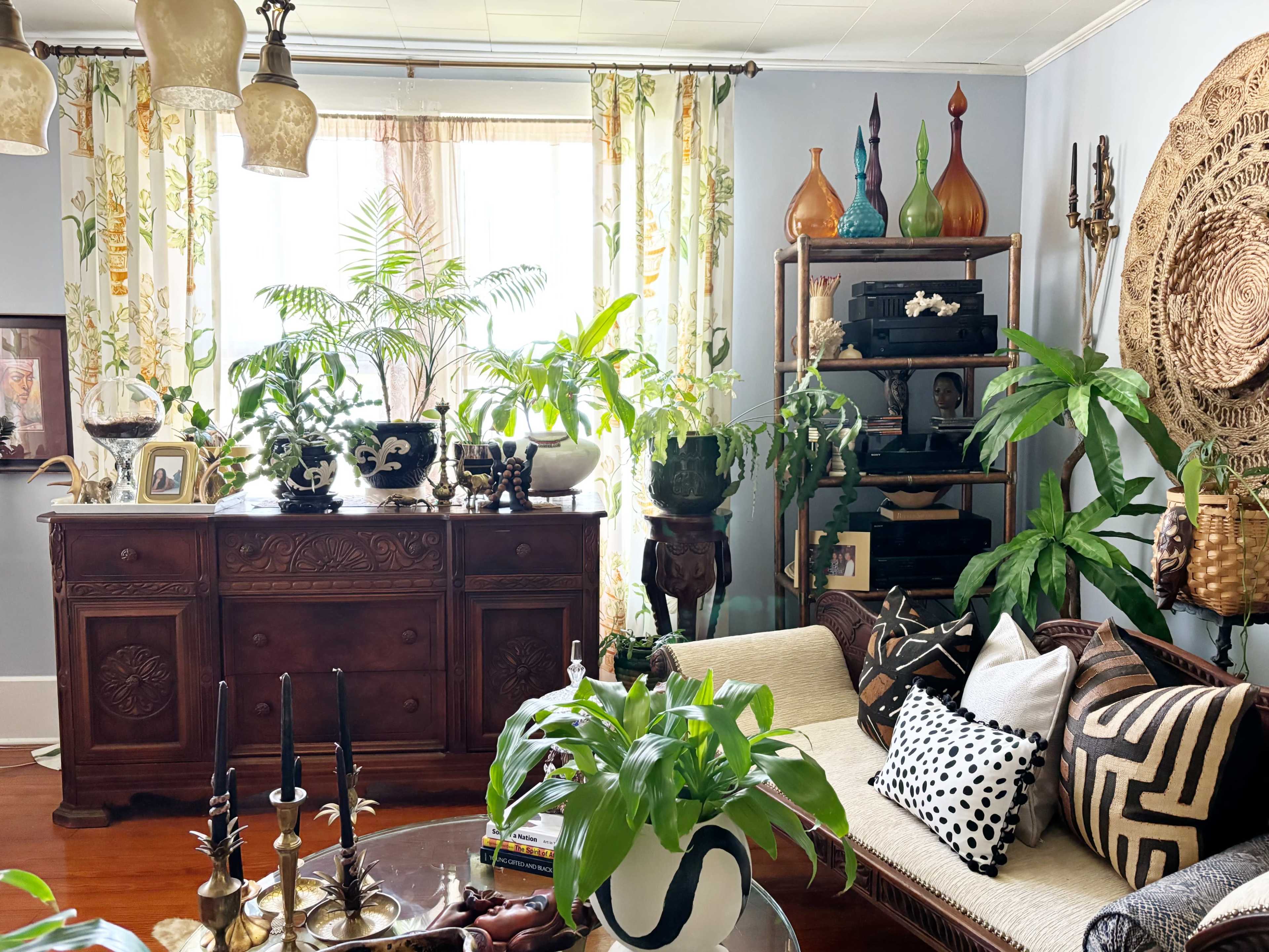 The image shows a cozy living room filled with various houseplants, an antique wooden sideboard, decorative glass vases, and a mix of patterned cushions on a sofa.