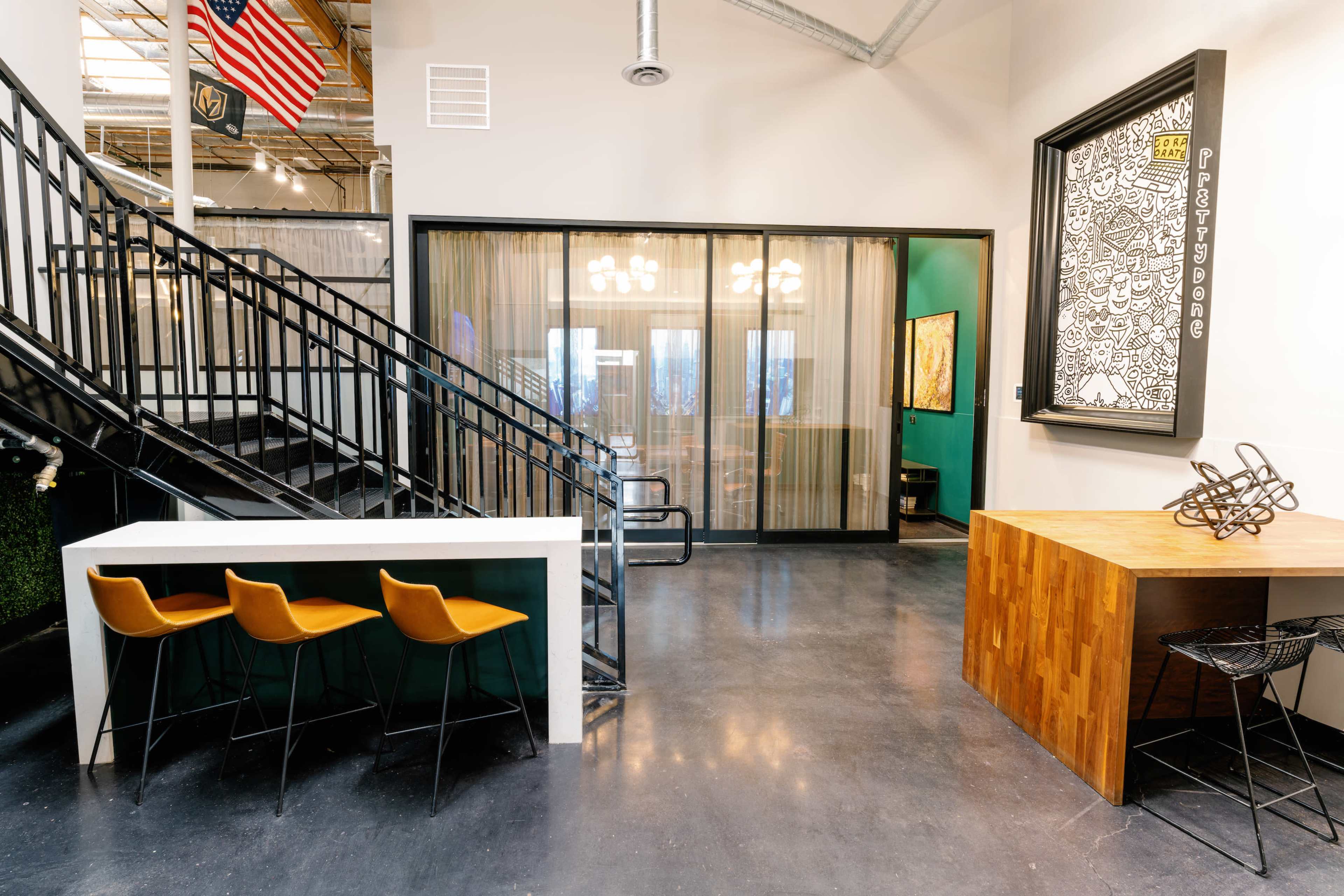 A modern office space featuring a staircase, seating area with bar stools, and a glass-enclosed meeting room.