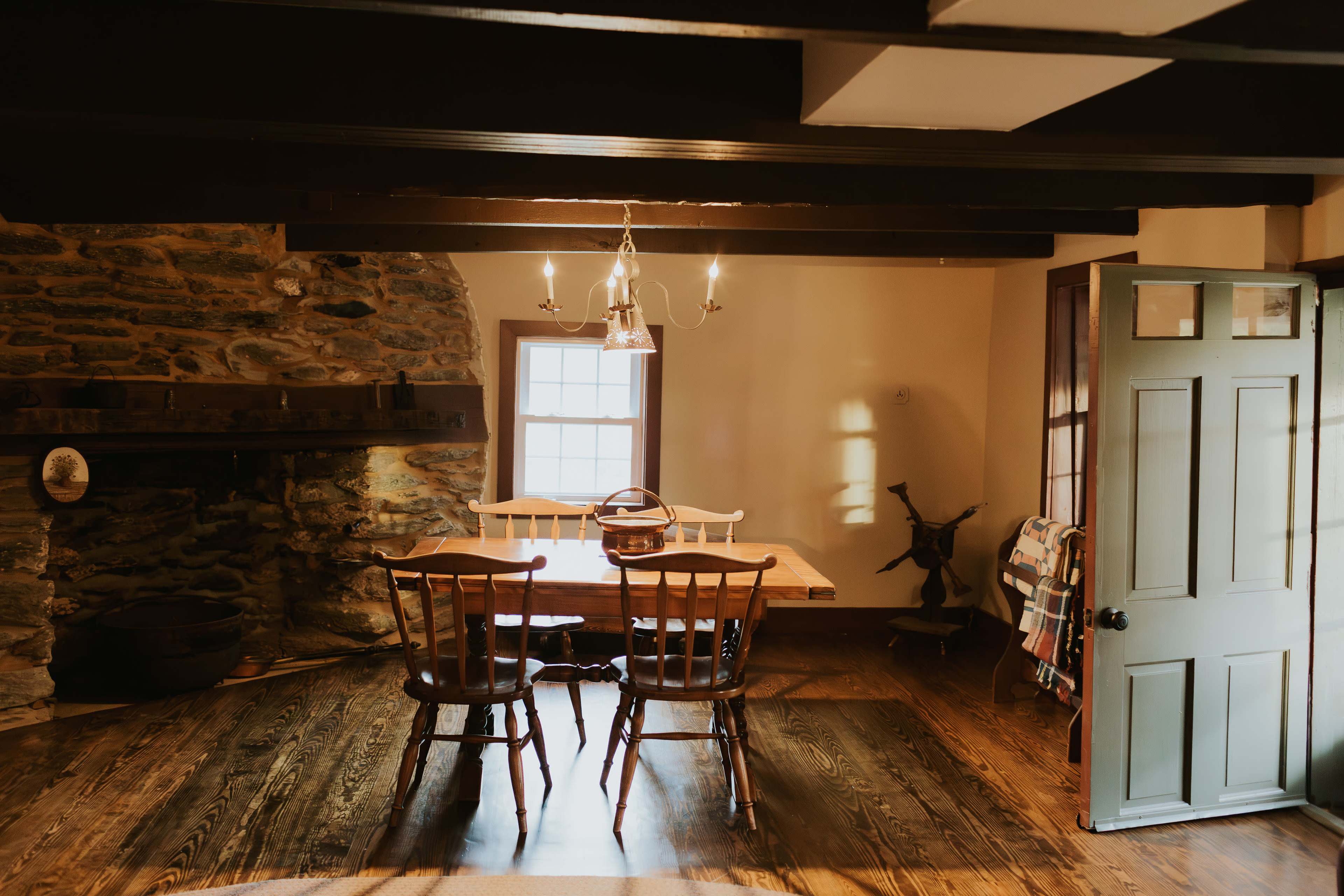 A rustic dining area with a wooden table, four chairs, and a stone wall, illuminated by natural light coming through a window.