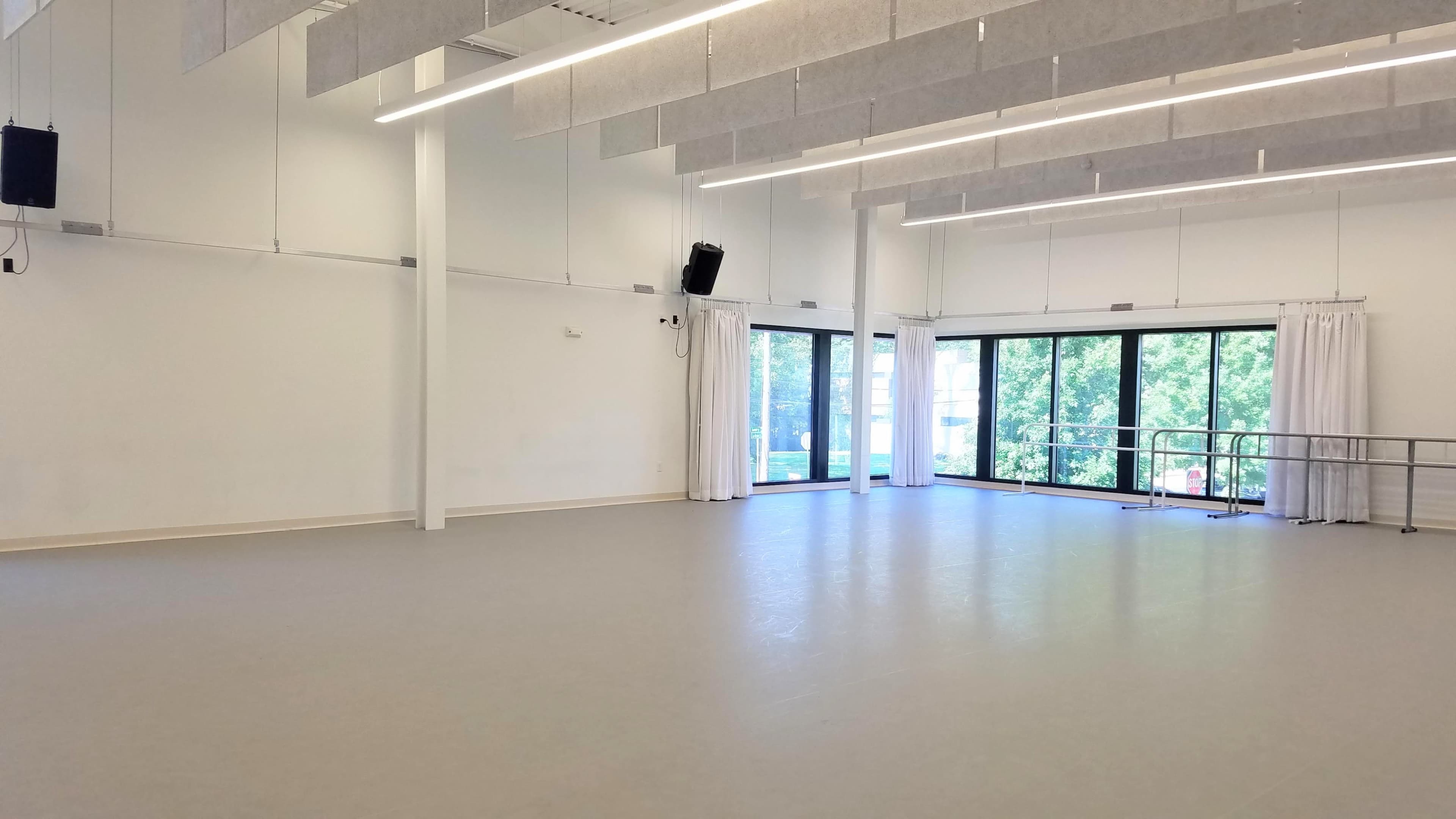 The image shows a spacious, well-lit dance studio with large windows and a mirrored wall.