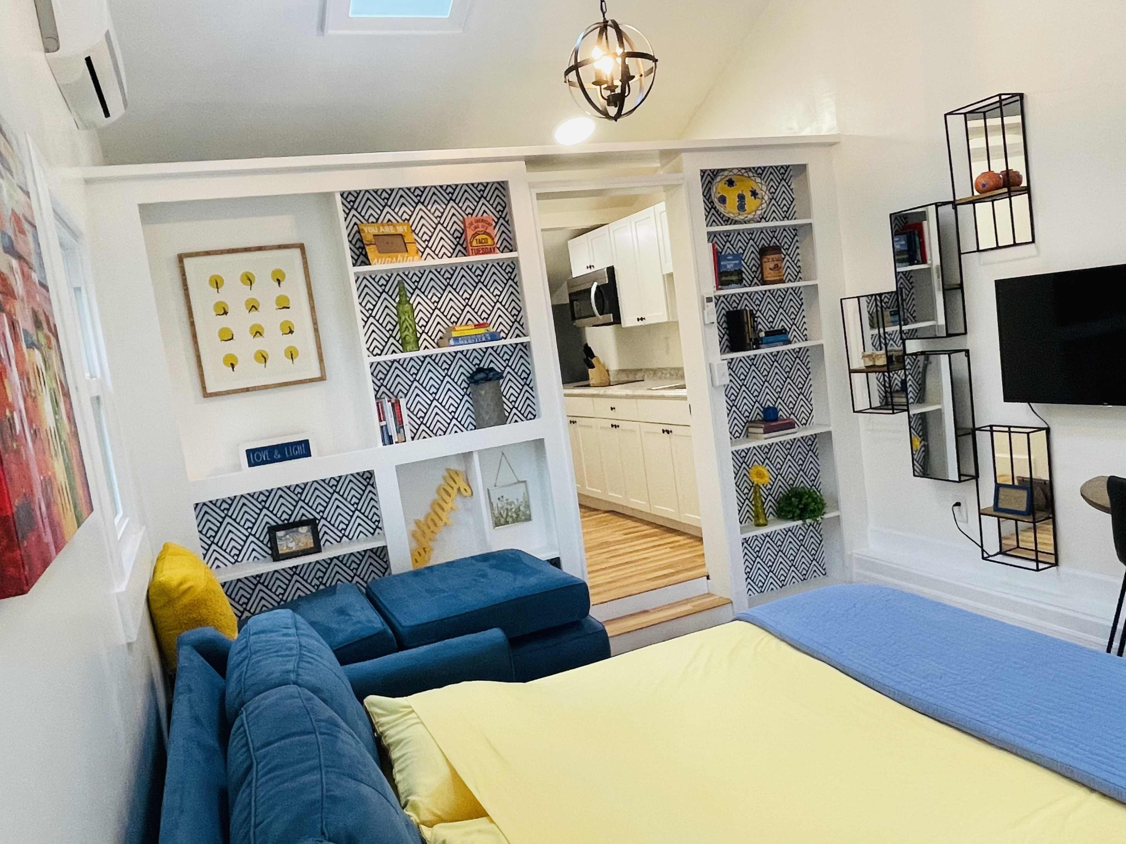 A living area with a blue couch, yellow bedding on a pull-out bed, and built-in shelves displaying decorative items and books, leading into a kitchen space.
