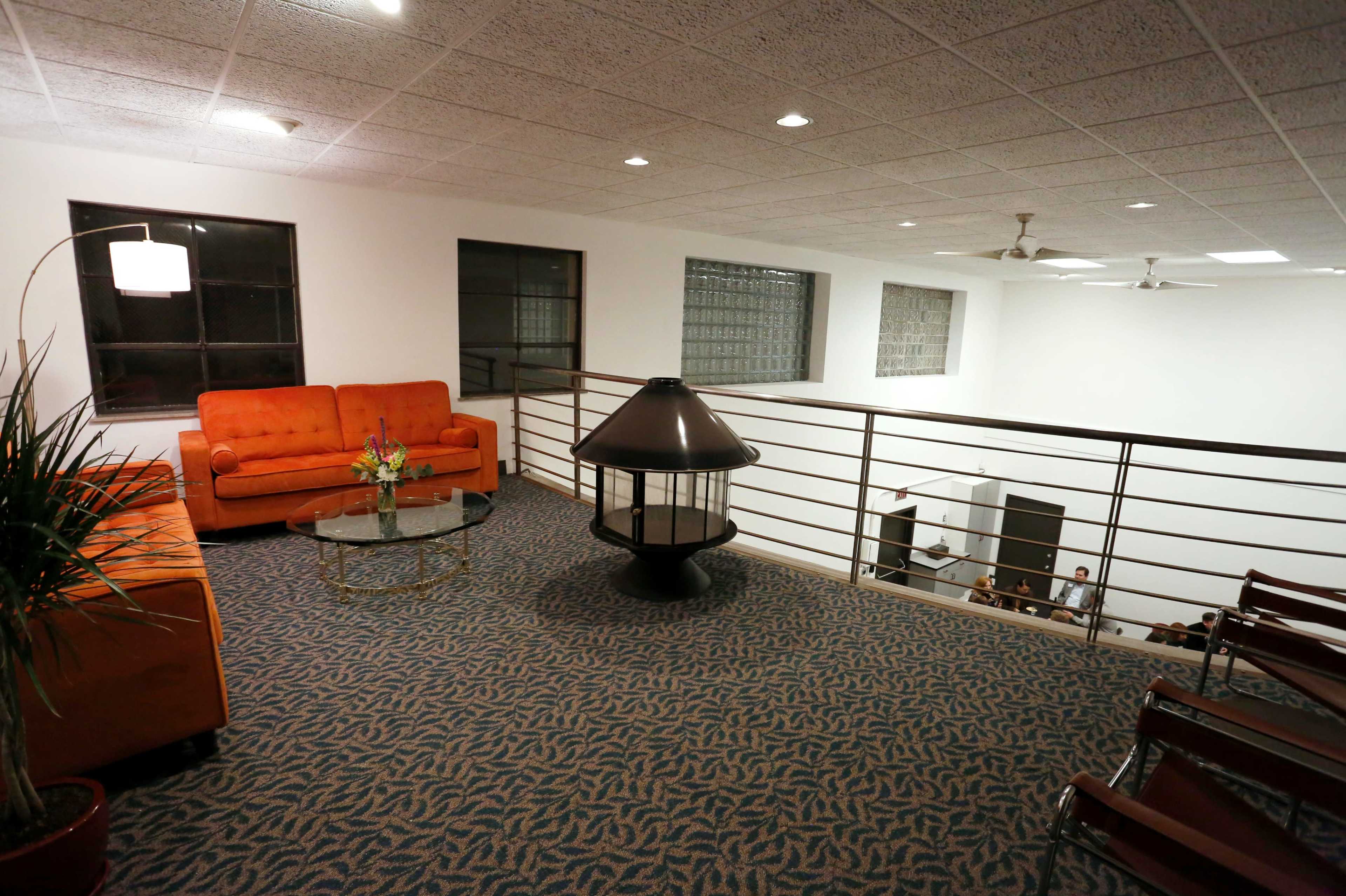 A spacious, well-lit mezzanine features an orange couch, a glass coffee table, and a lamp, overlooking a room with chairs and people.