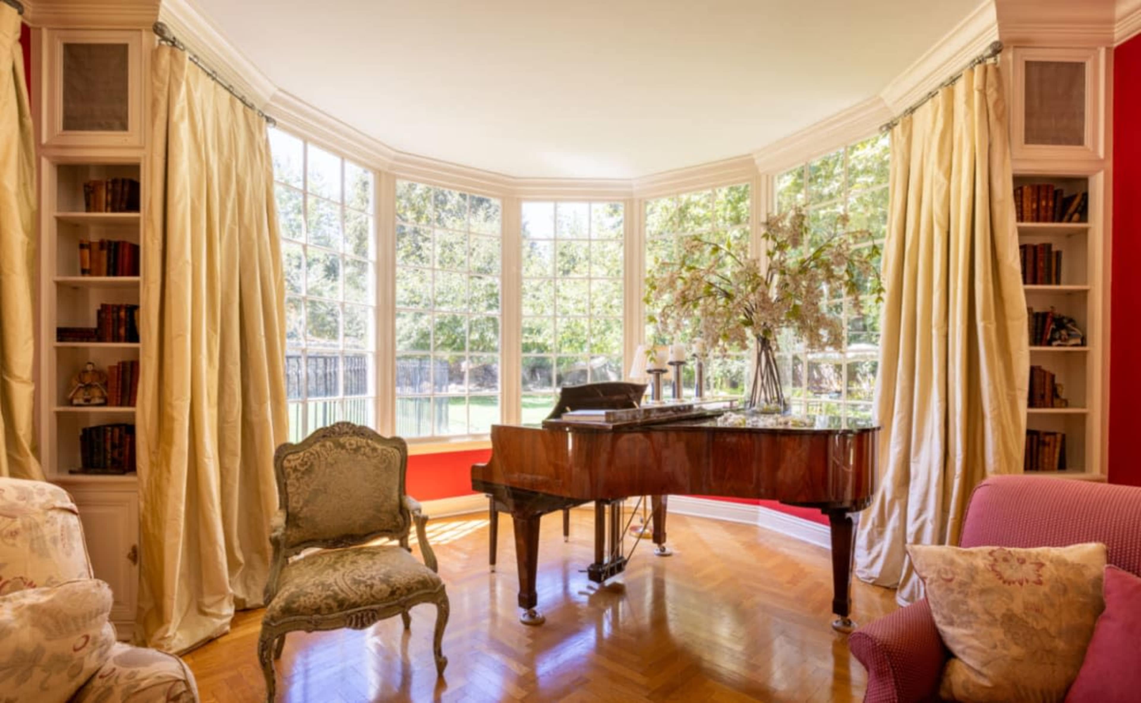 A grand piano sits beside large windows adorned with light curtains, surrounded by bookshelves and elegant seating in a well-lit room.