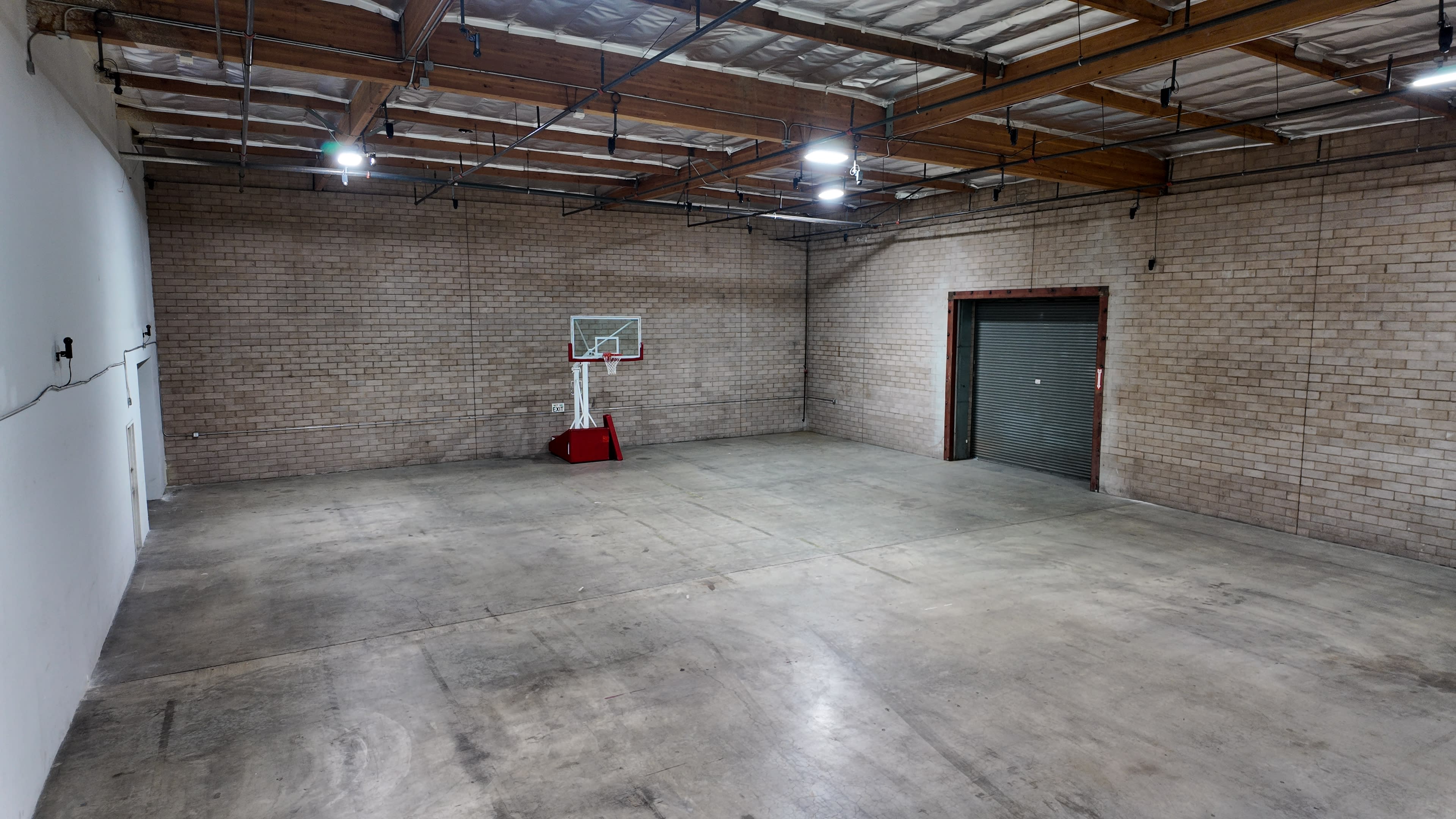 The image shows an empty indoor basketball court with a hoop on one side and a large roll-up door on the opposite wall.