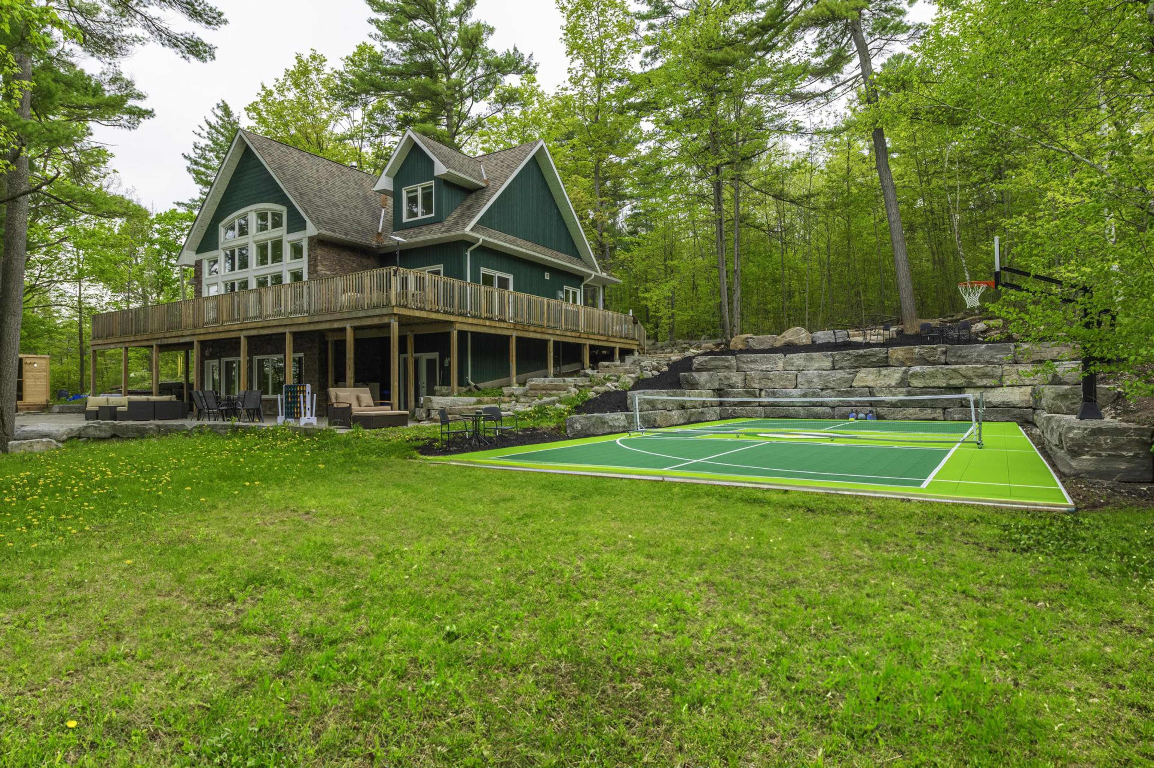 A large green house with a deck overlooks a basketball court situated on a landscaped yard surrounded by trees.