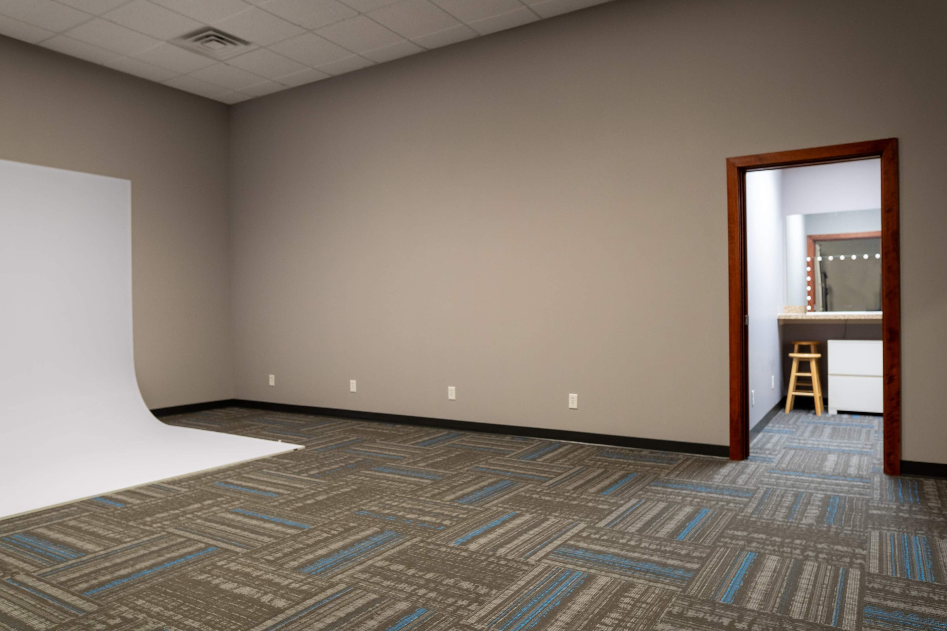 The image shows an empty room with a white backdrop on one side, a door leading to another room, and a mirror with lights visible through the doorway.