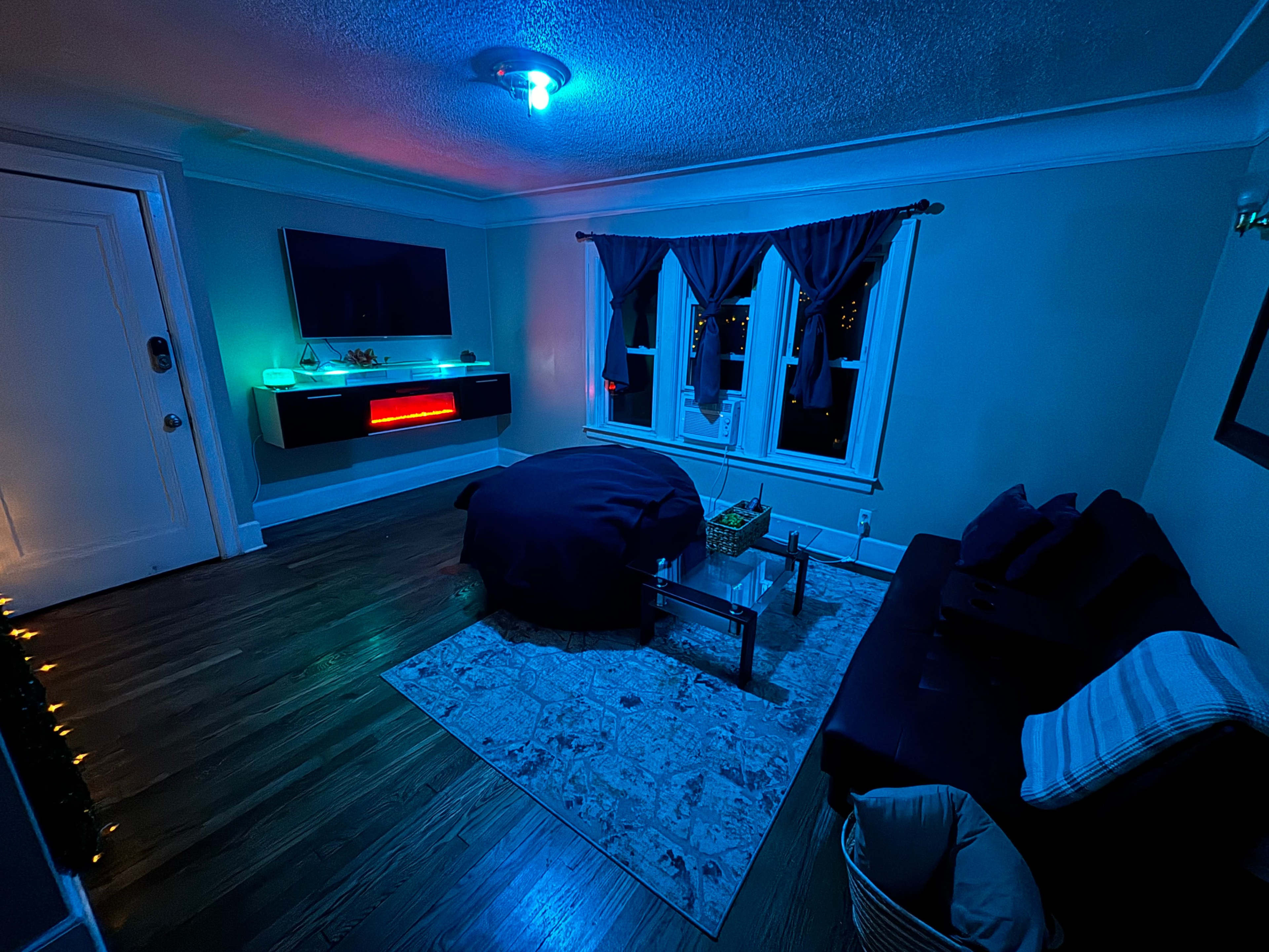 The room features a dark theme with blue lighting, a large television on the wall, a bed in the center, and a seating area with a rug.
