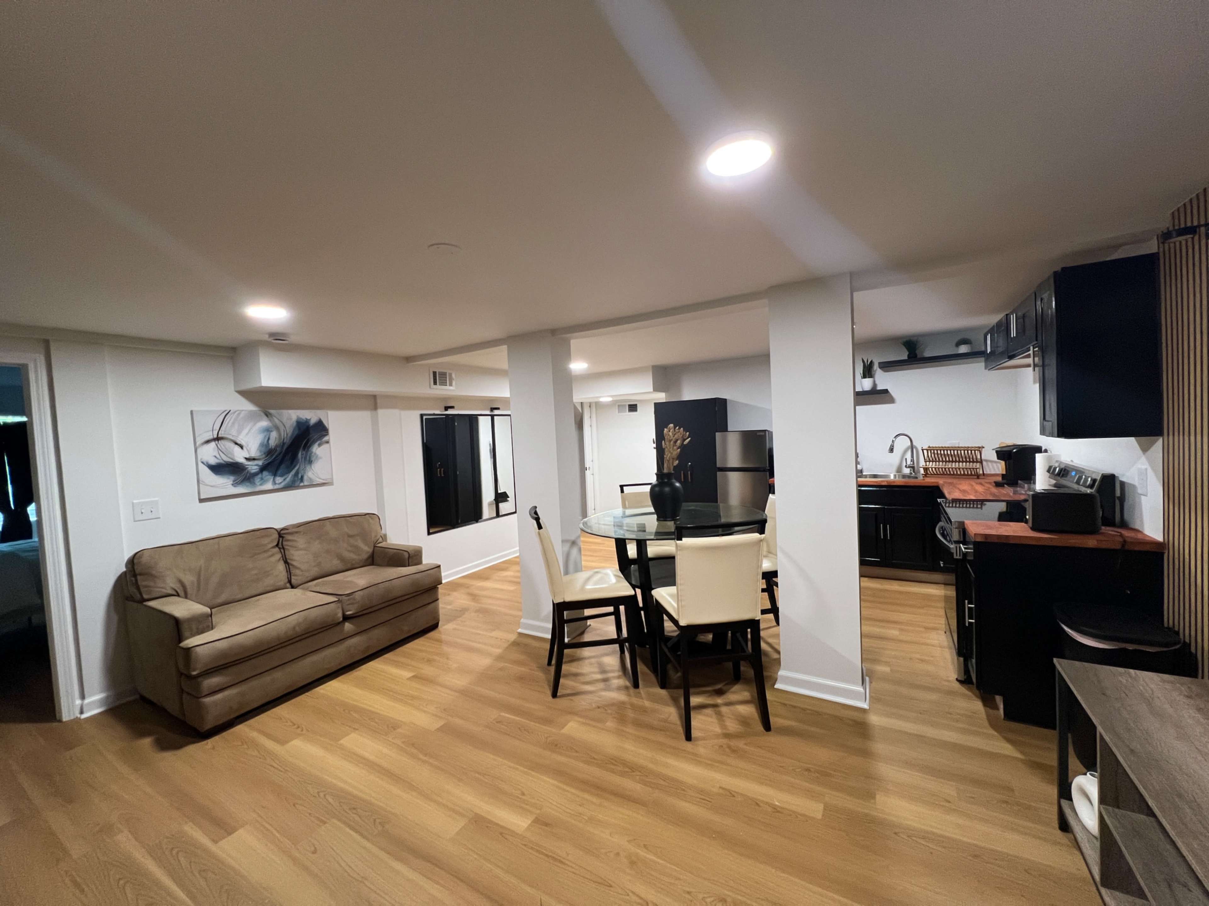 The image shows a modern, open-concept living space featuring a beige couch, a dining table with chairs, and a kitchen area with black cabinets and appliances.