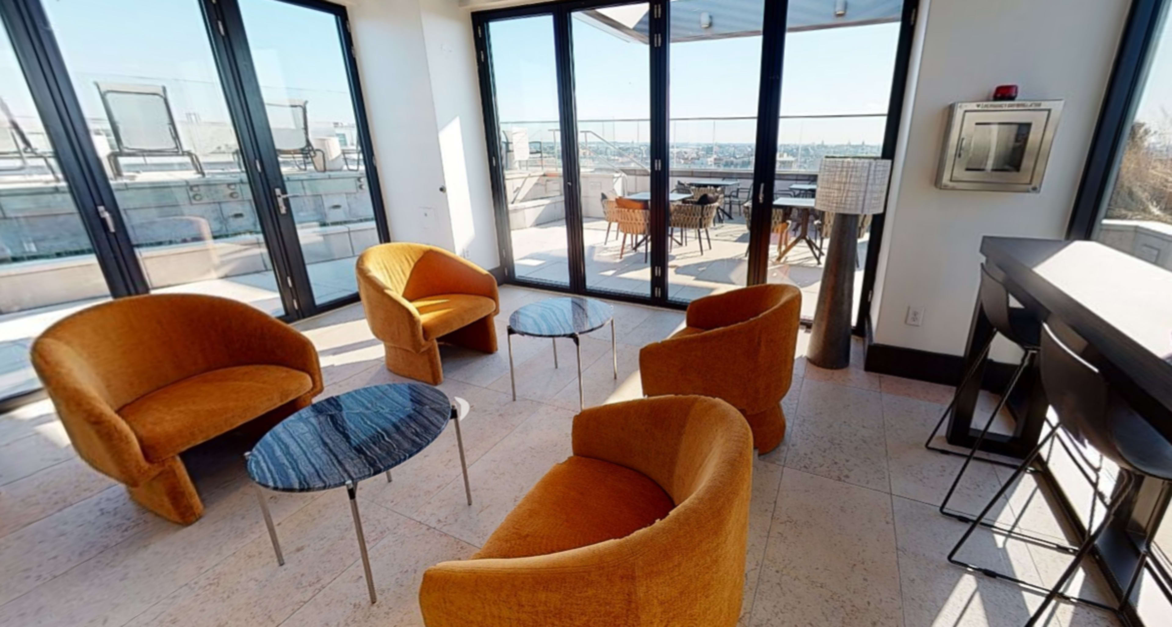 A modern lounge area with four orange chairs and two round black tables, featuring large windows that overlook a city skyline.