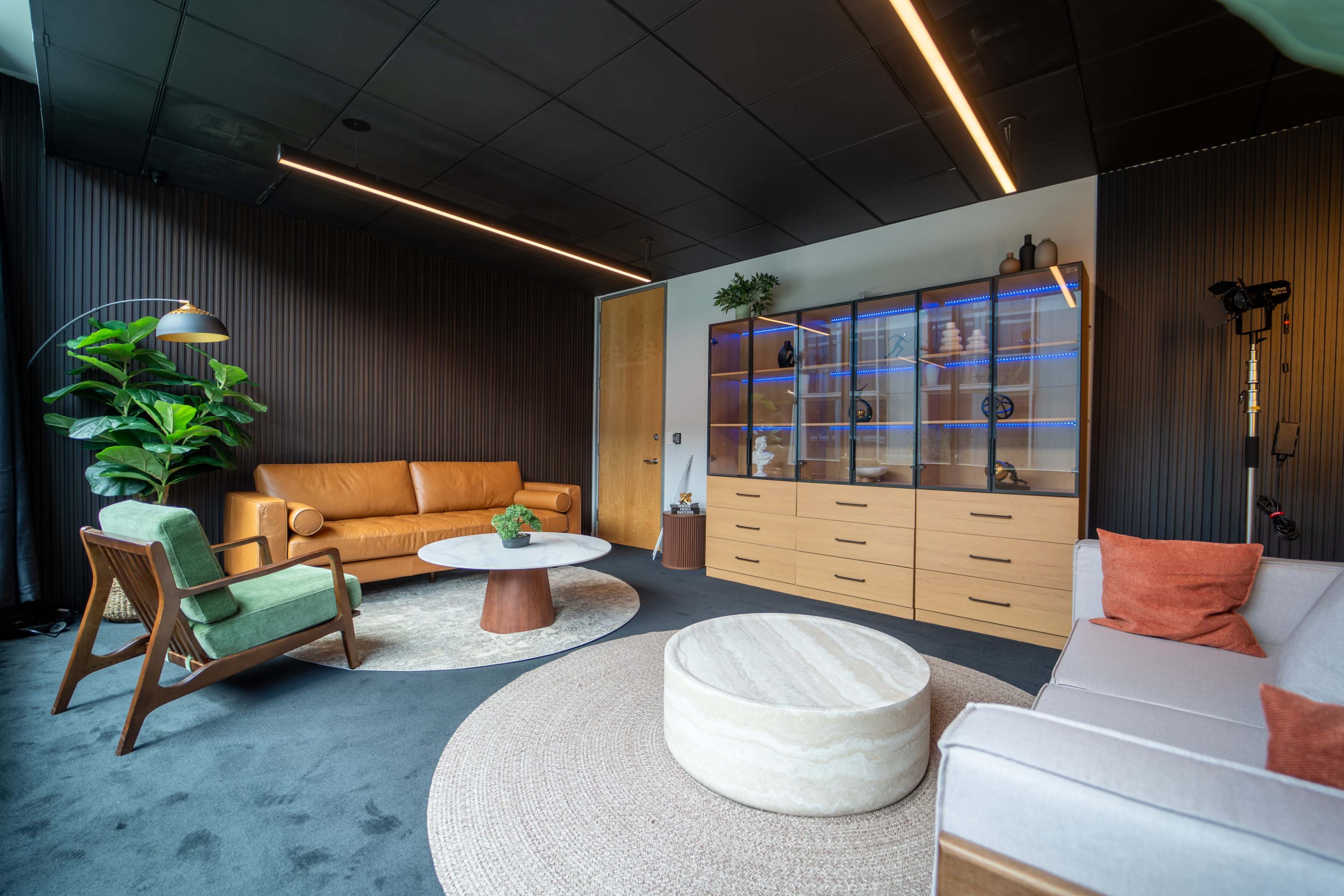 The image showcases a modern interior lounge with a mix of leather and fabric seating, a round coffee table, and a glass-fronted cabinet illuminated with blue lighting.