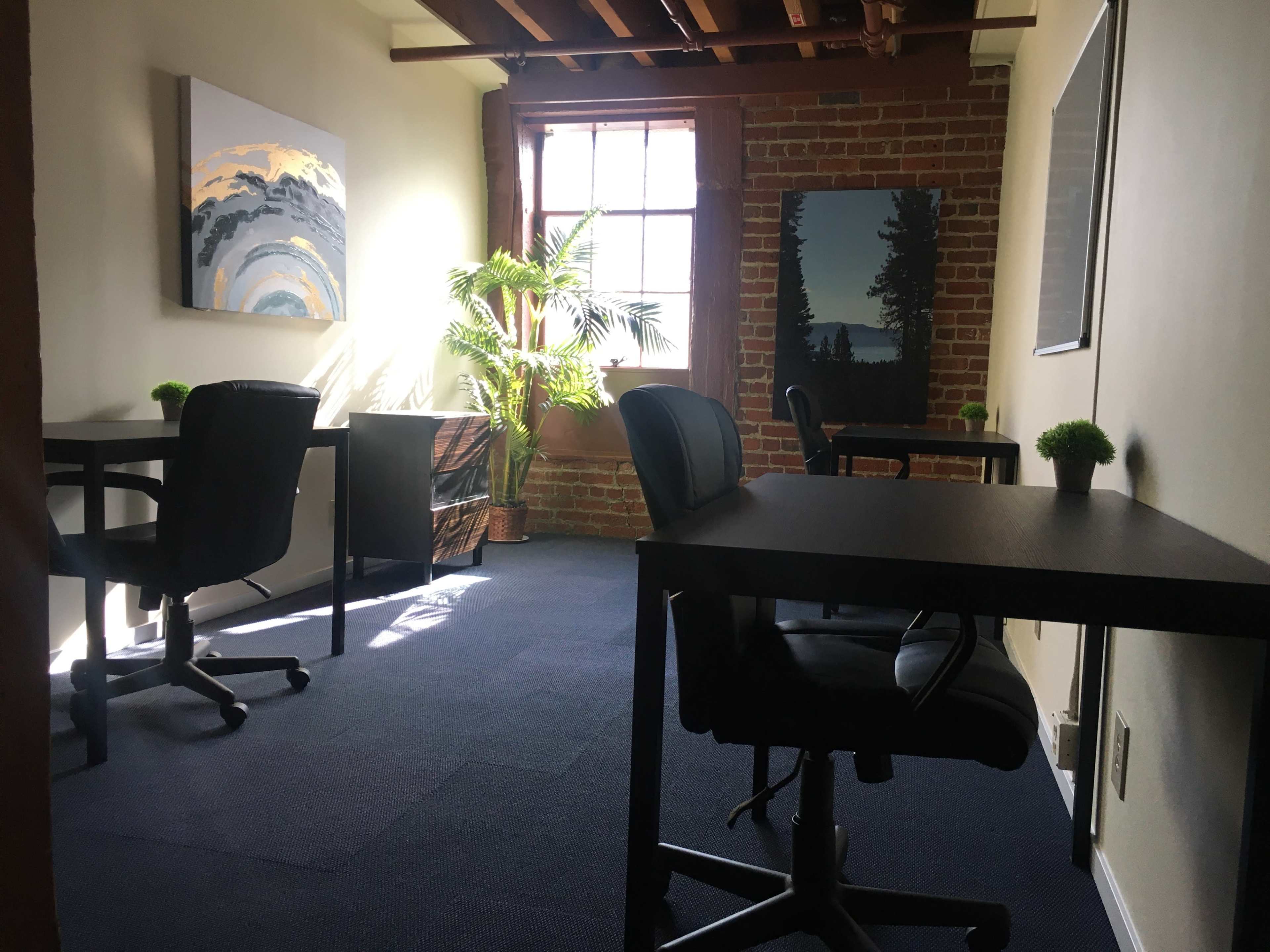 The image shows an office space with two empty black desks, chairs, and decor including plants and artwork, illuminated by natural light from a window.
