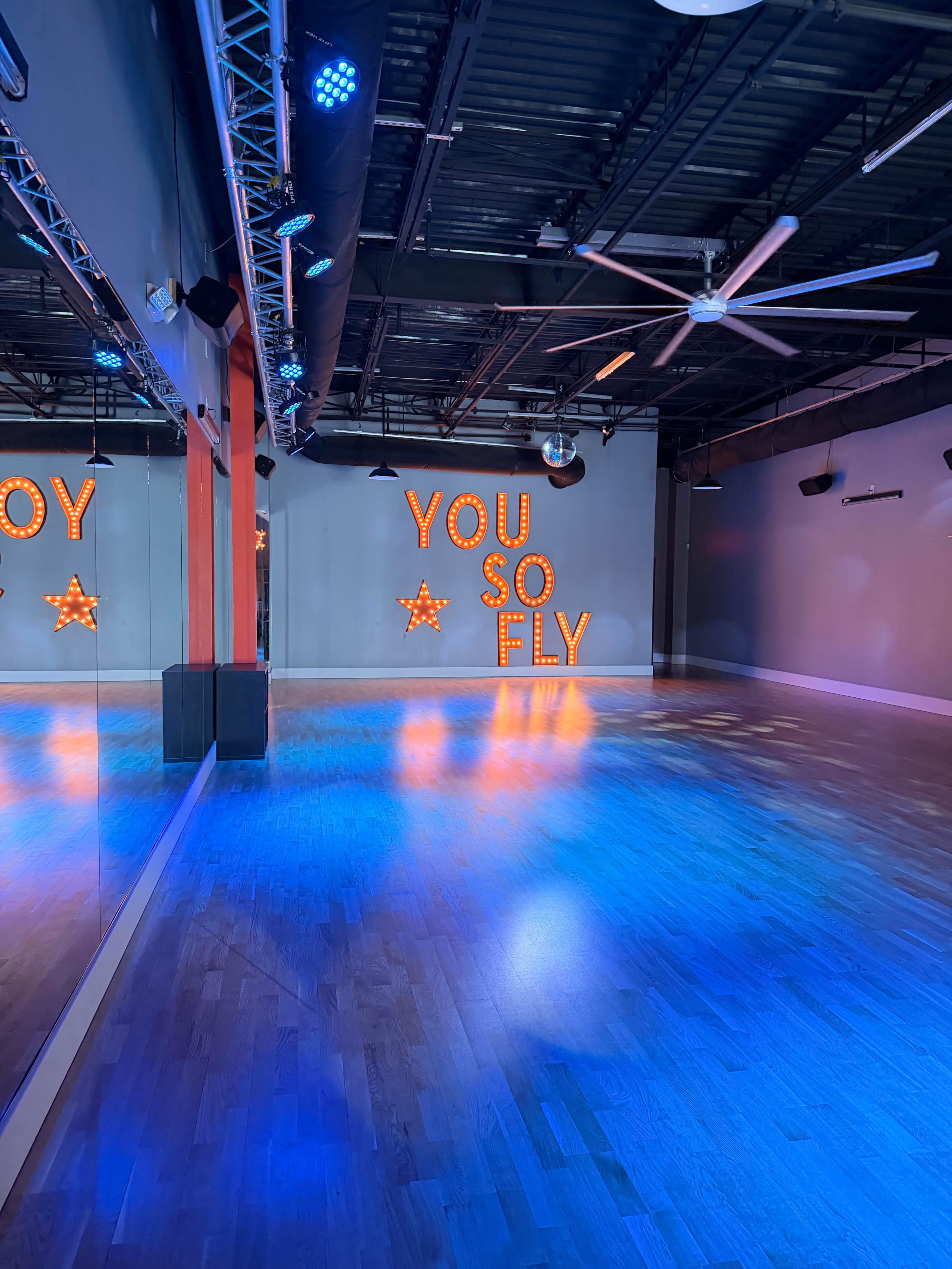 The image shows a spacious dance studio with wooden floors, illuminated by blue and orange lights, featuring a wall with the illuminated words "YOU SO FLY" and a star decoration.