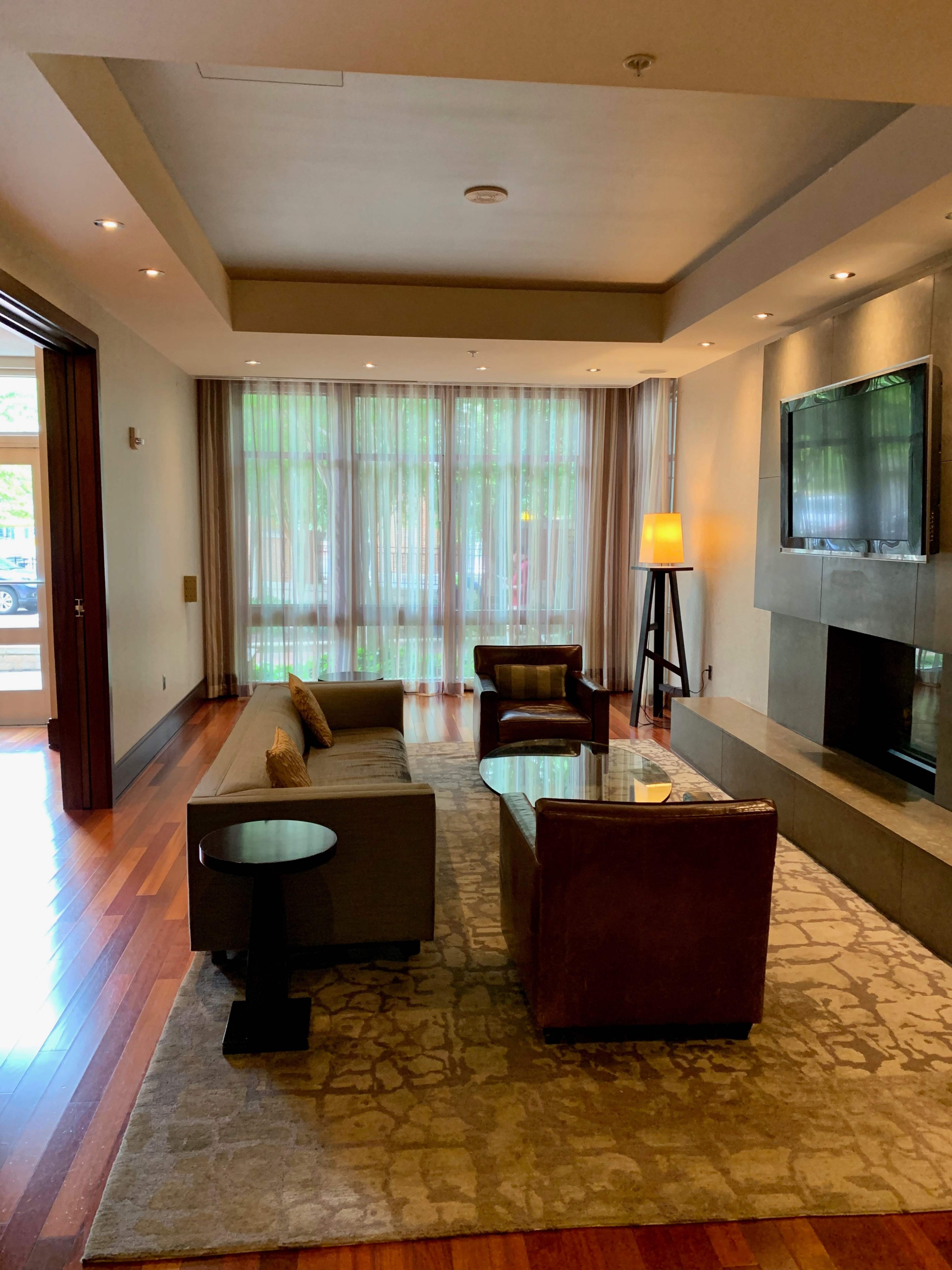 A modern living room features a sofa, an armchair, a coffee table, and a lamp, with large windows covered by sheer curtains allowing natural light to fill the space.