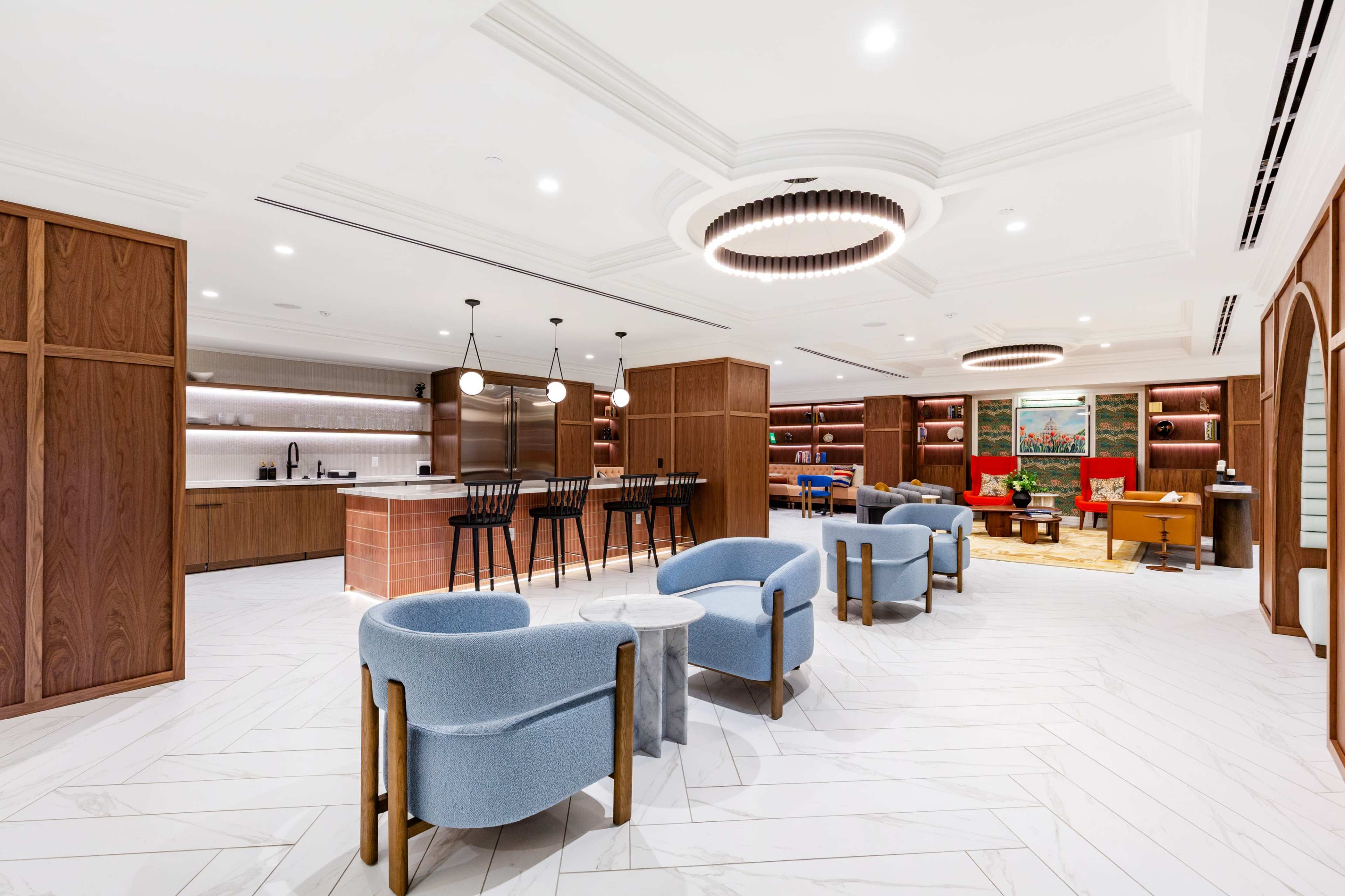 The image shows a modern lounge area with blue chairs and a kitchen bar, featuring warm wood accents and ambient lighting.