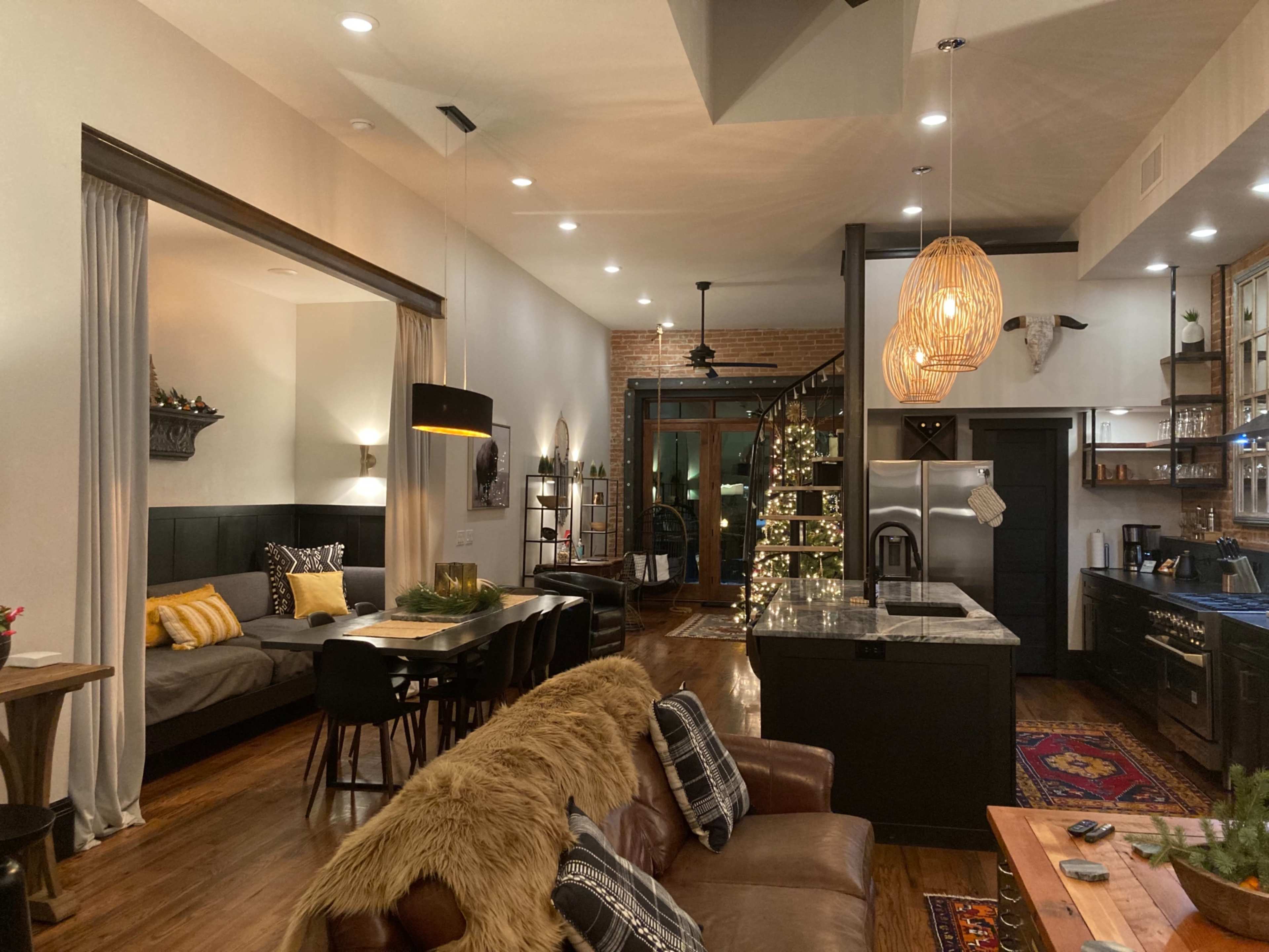 The image shows a modern open-concept living space featuring a kitchen, dining area, and a cozy sitting area adjacent to a decorative Christmas tree.