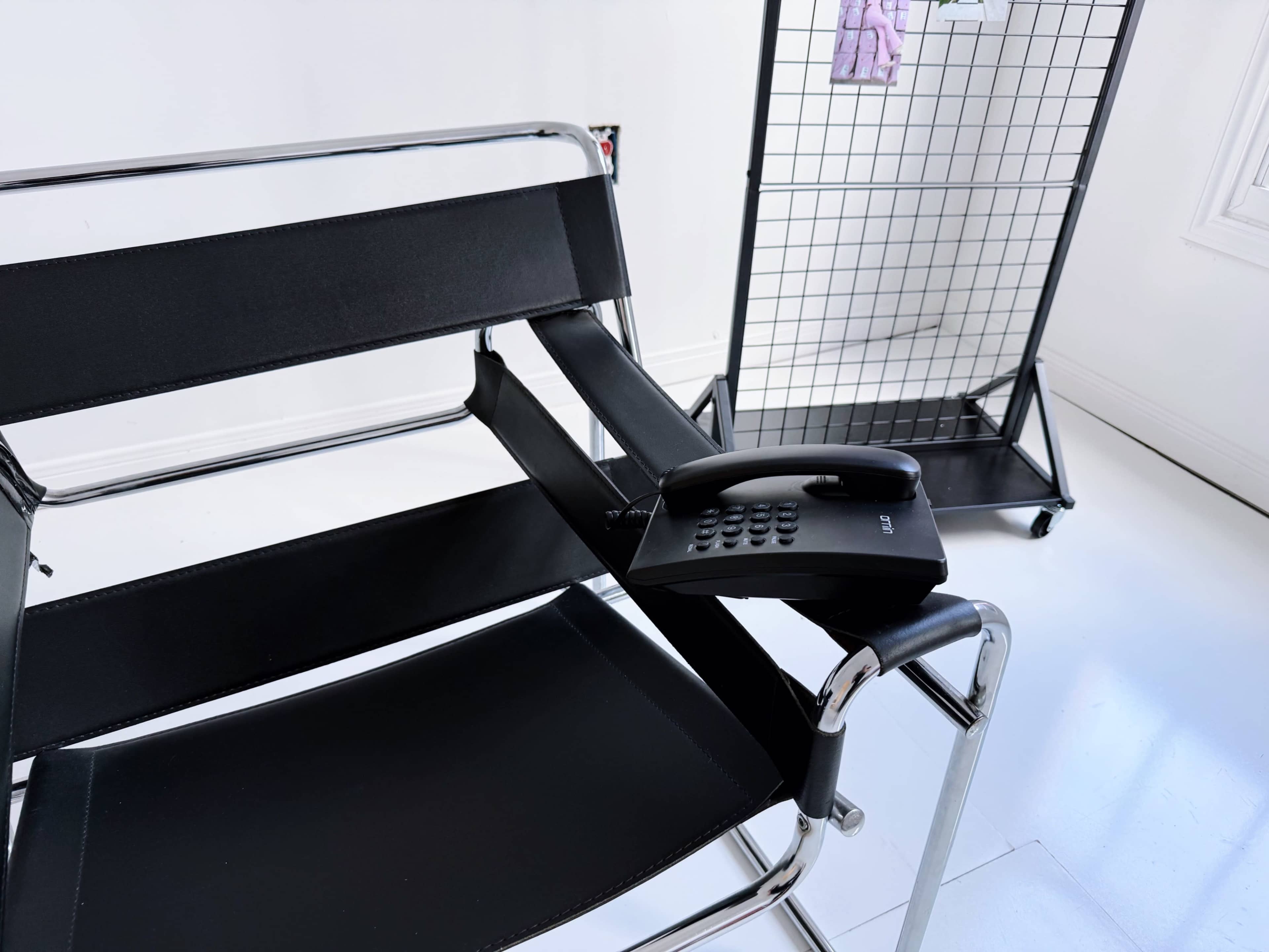A black metal-framed chair with fabric seating has a telephone placed on its armrest, alongside a black grid-style clothing rack in the background.