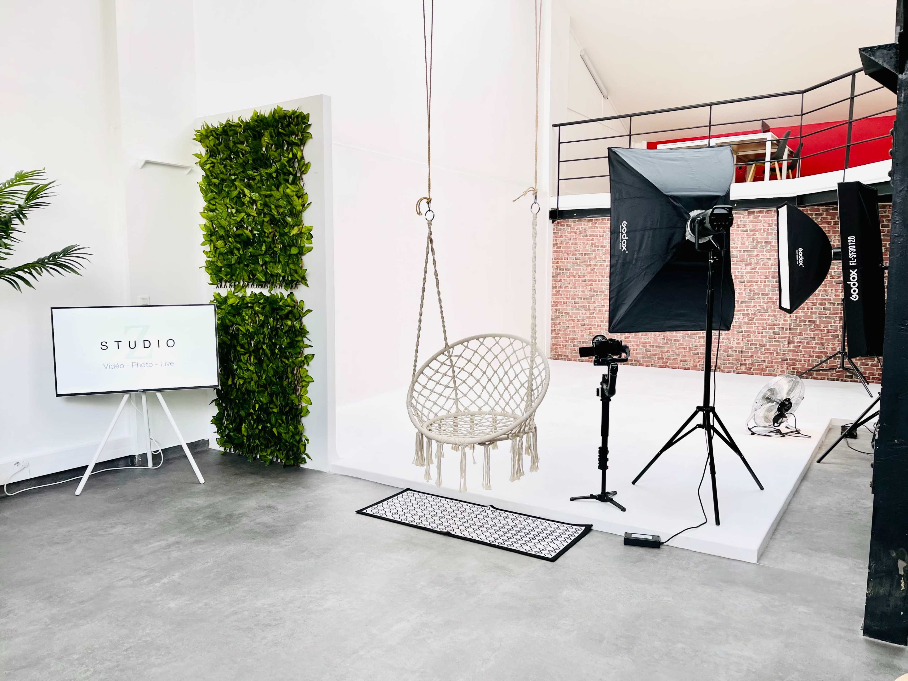 A minimalistic studio space featuring a hanging chair, a backdrop of greenery, and equipment for photography, including lights and a camera on a tripod.