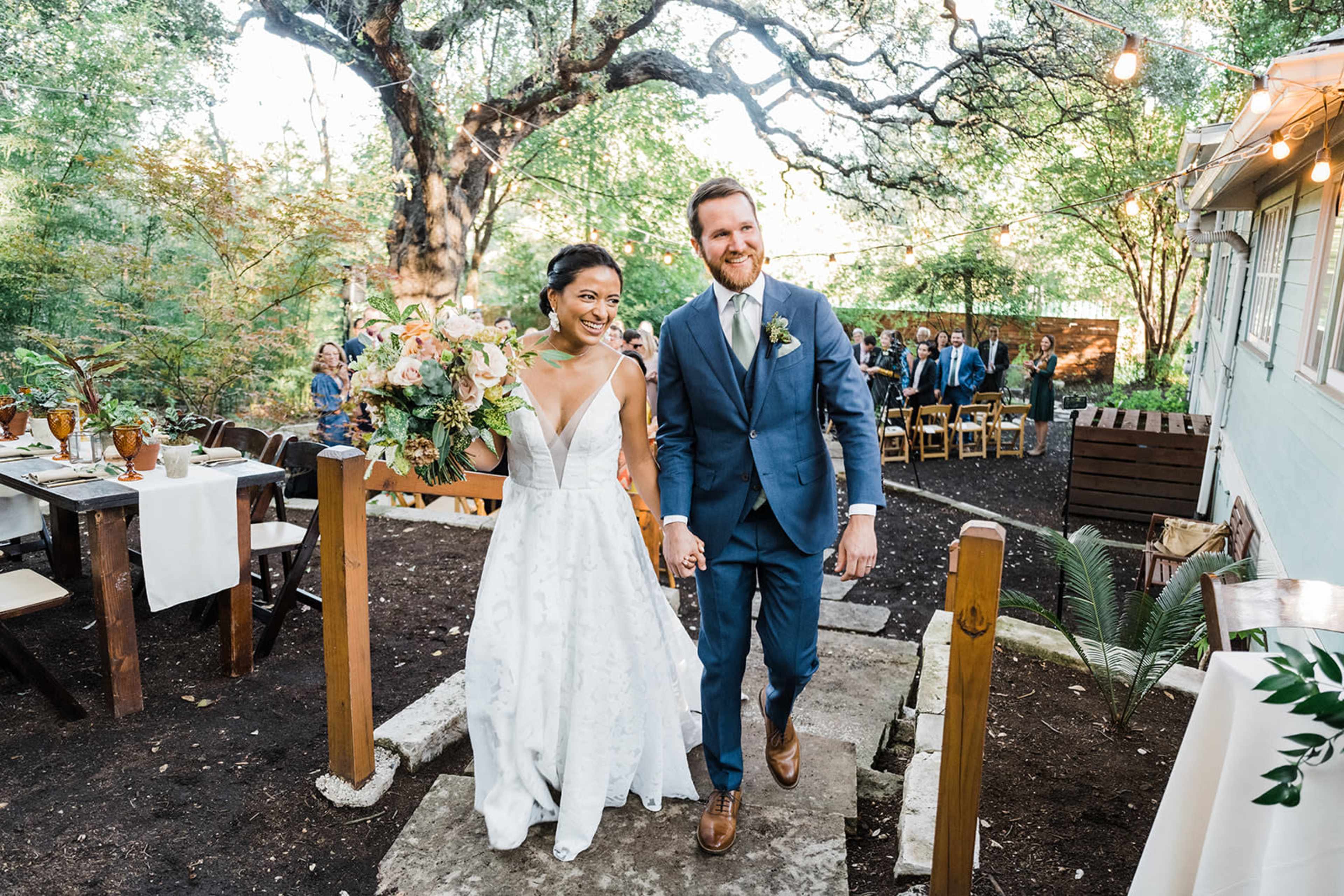 A bride and groom walk hand-in-hand down a stone path, smiling, as they exit a wedding ceremony in a garden setting.