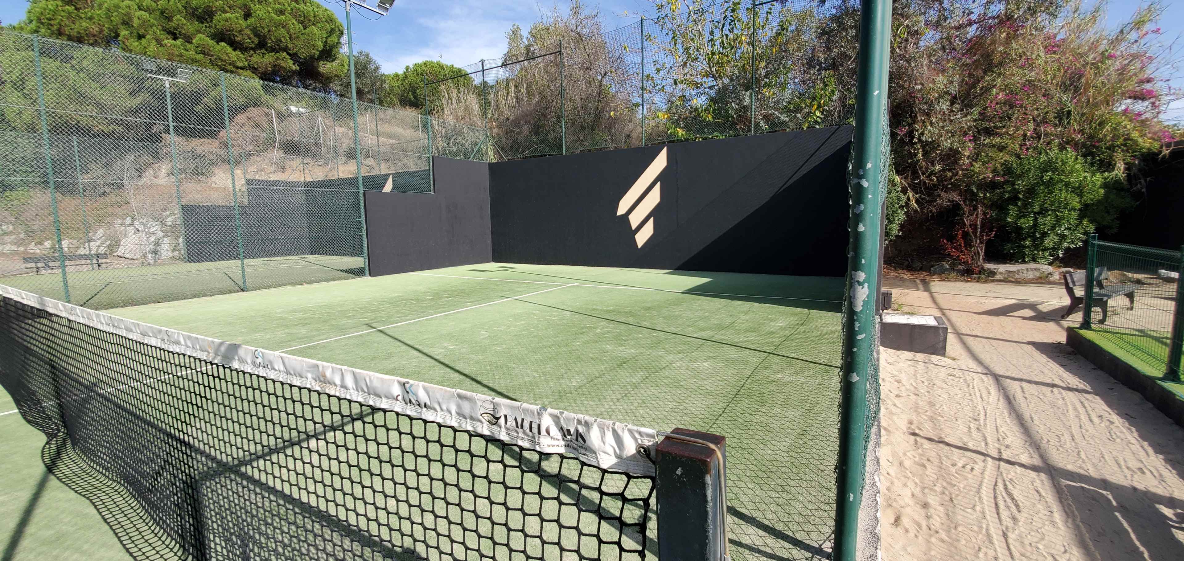 A green artificial turf tennis court enclosed with a black wall and a sandy pathway nearby.
