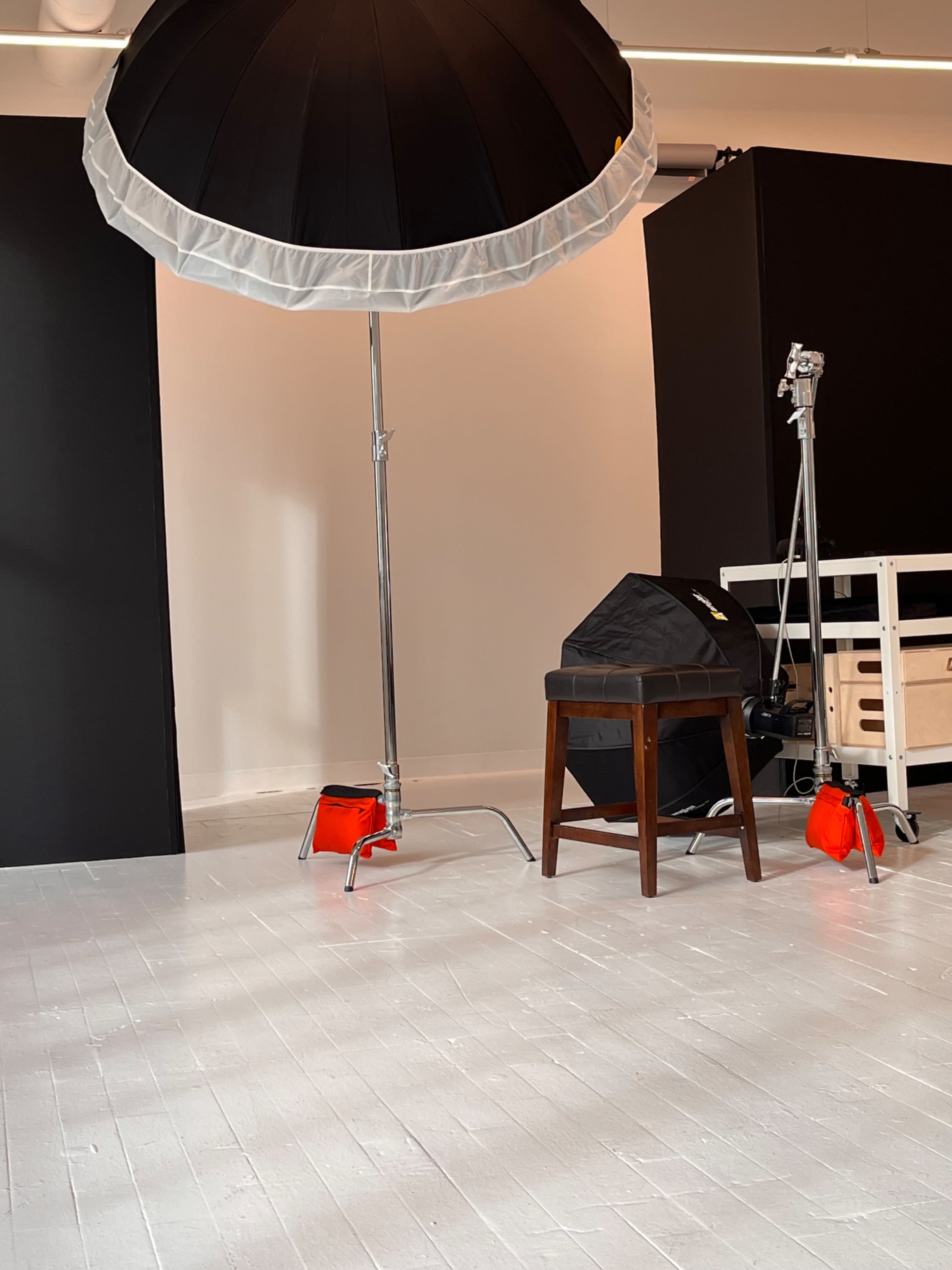 A photography studio setup with a large umbrella light, a wooden stool, and various equipment against a white floor.