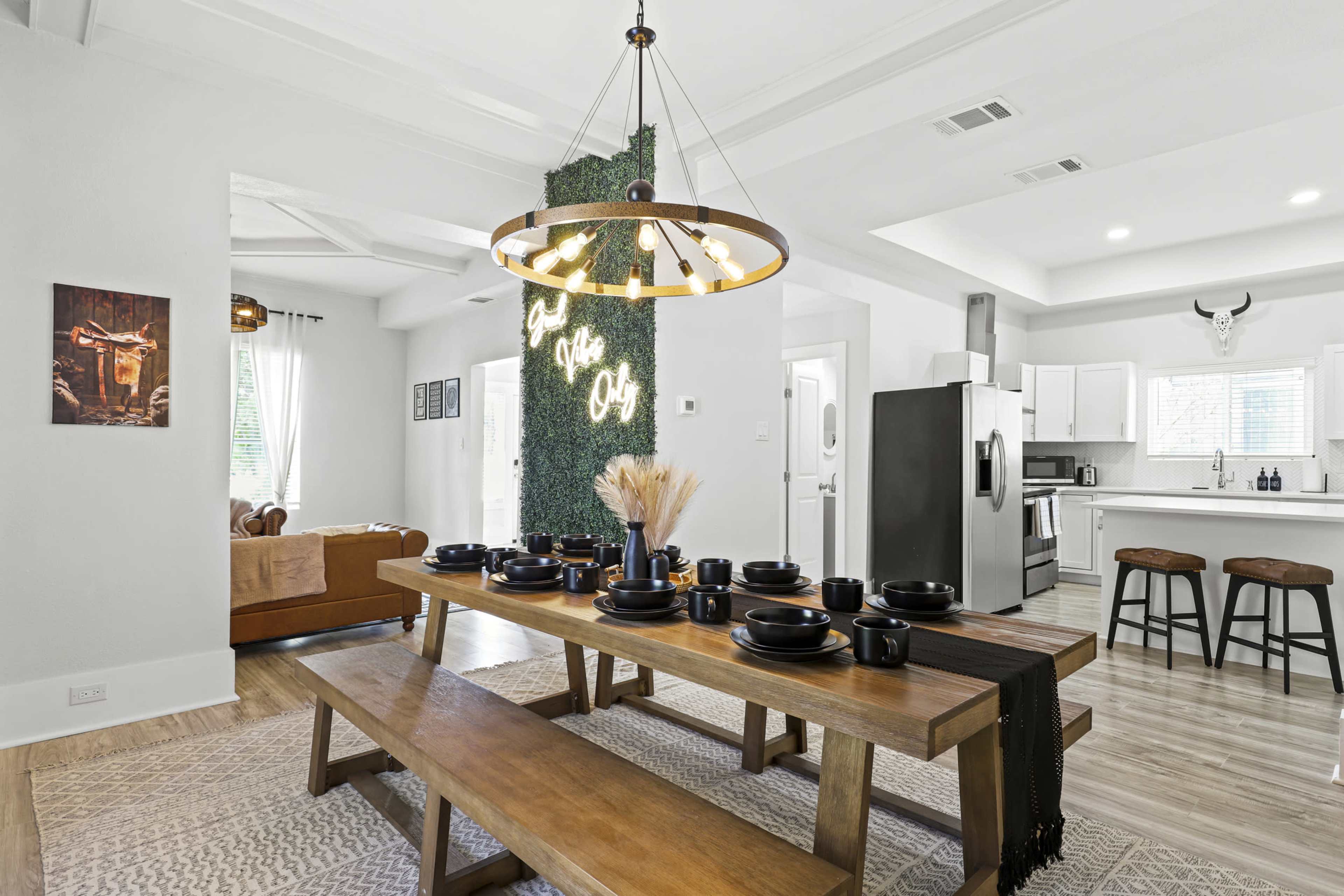 A spacious dining area features a large wooden table set for a meal, with a modern chandelier above and a kitchen visible in the background.